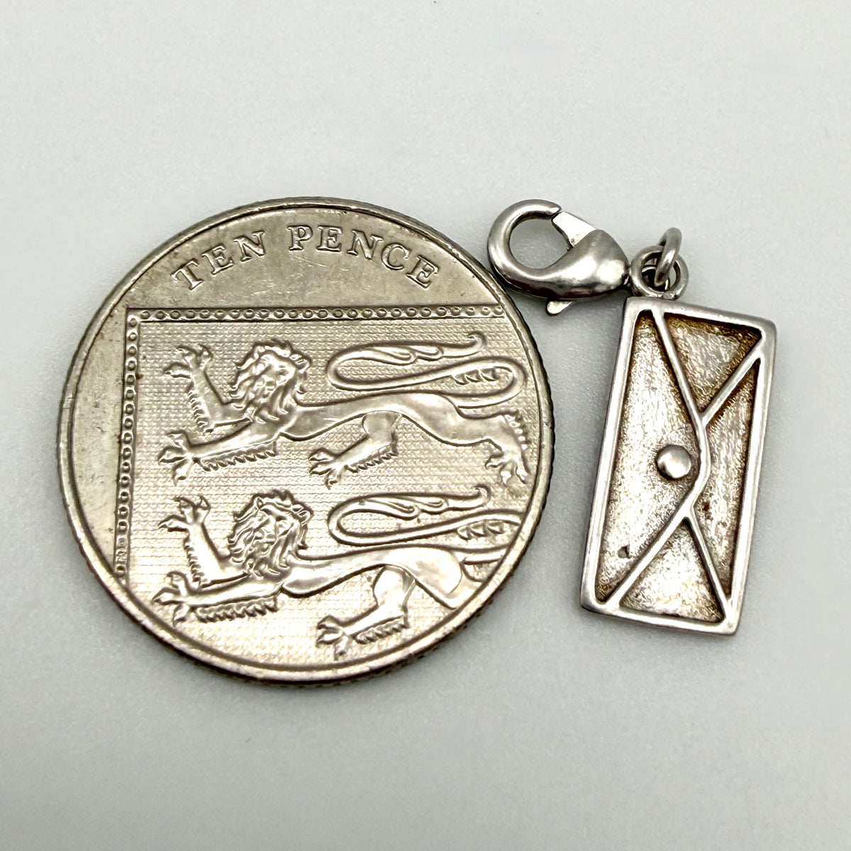 A sterling silver charm shaped like a vintage British ten pence coin next to a matching rectangular envelope pendant, displayed on a light gray background.