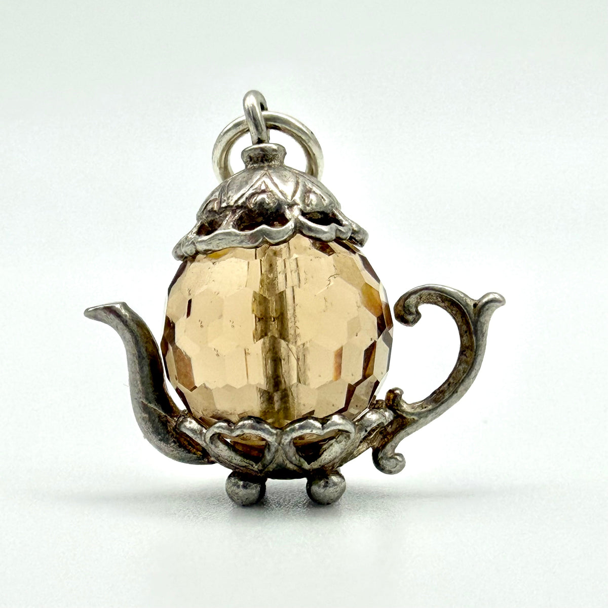 A vintage-style teapot pendant charm featuring a faceted amber-yellow glass sphere set in an ornate silver-tone metal frame, shown on a plain light background.