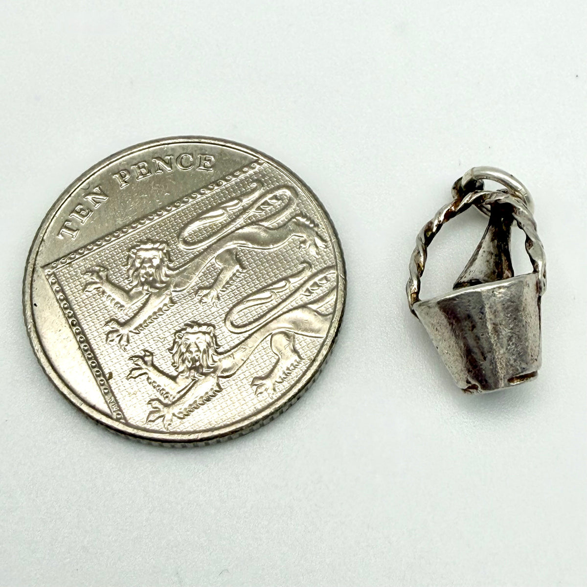 A small silver pendant or charm shown next to a UK ten-pence coin for scale, on a plain light background.