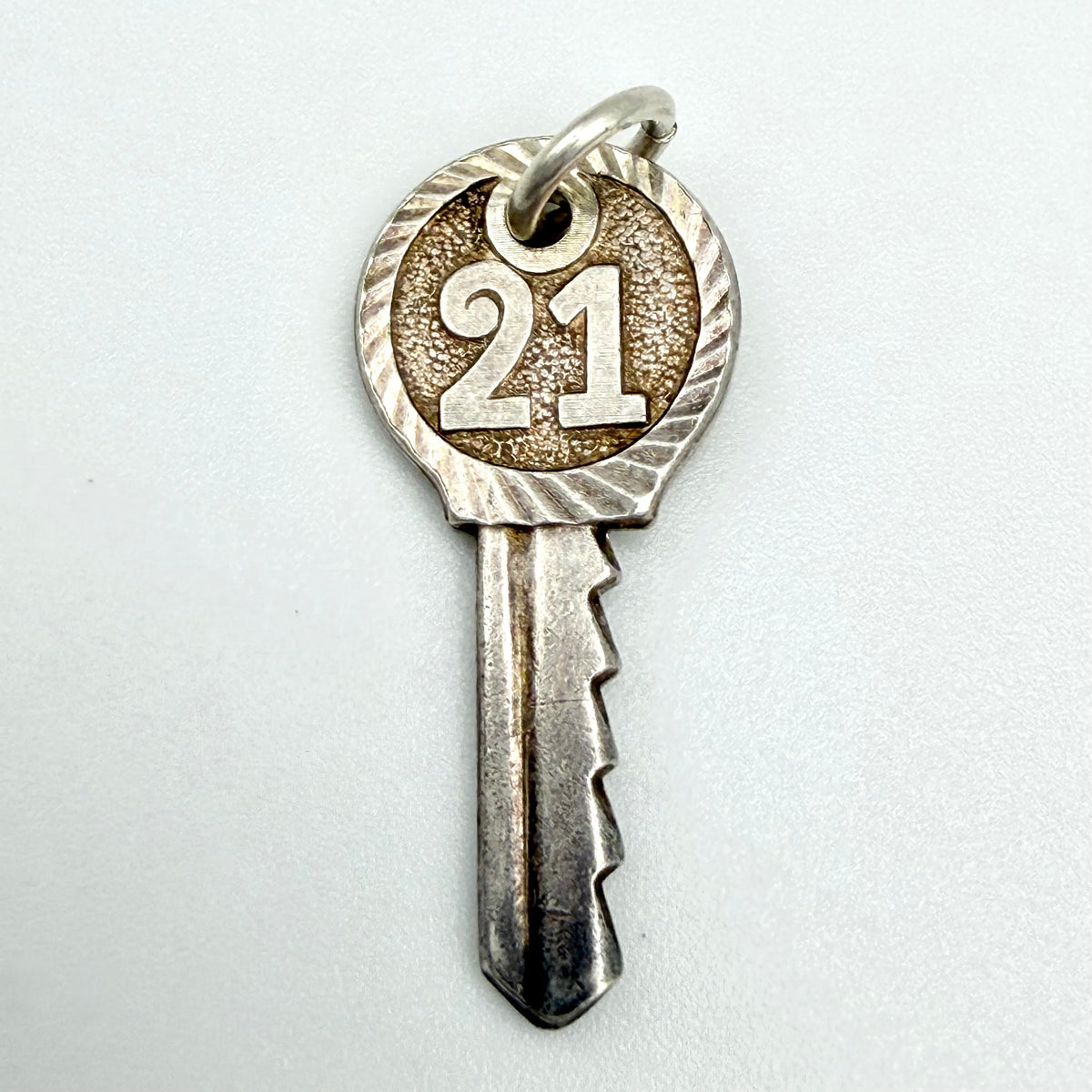 A silver-colored metal key-shaped pendant charm featuring the engraved number '21' on a textured circular head with a decorative wavy border, shown on a plain light background.