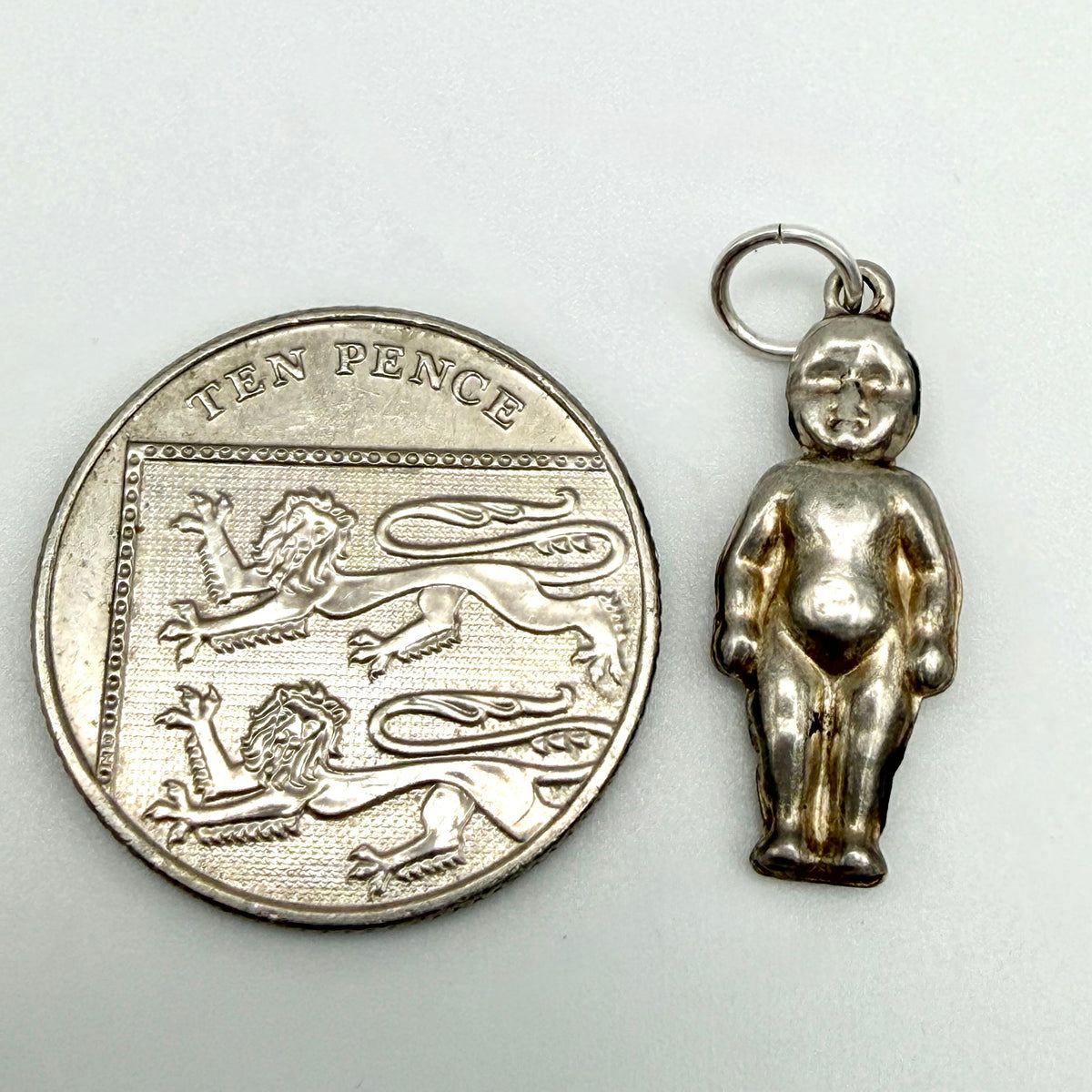 A small silver figurine charm of a standing nude child, shown next to a silver ten pence coin for scale on a plain light background.