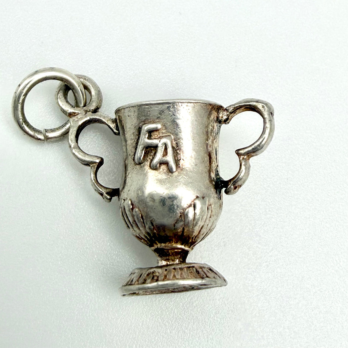 A silver trophy cup pendant charm with two handles, a small loop at the top, and the letters 'FA' engraved on the front, shown on a plain light background.
