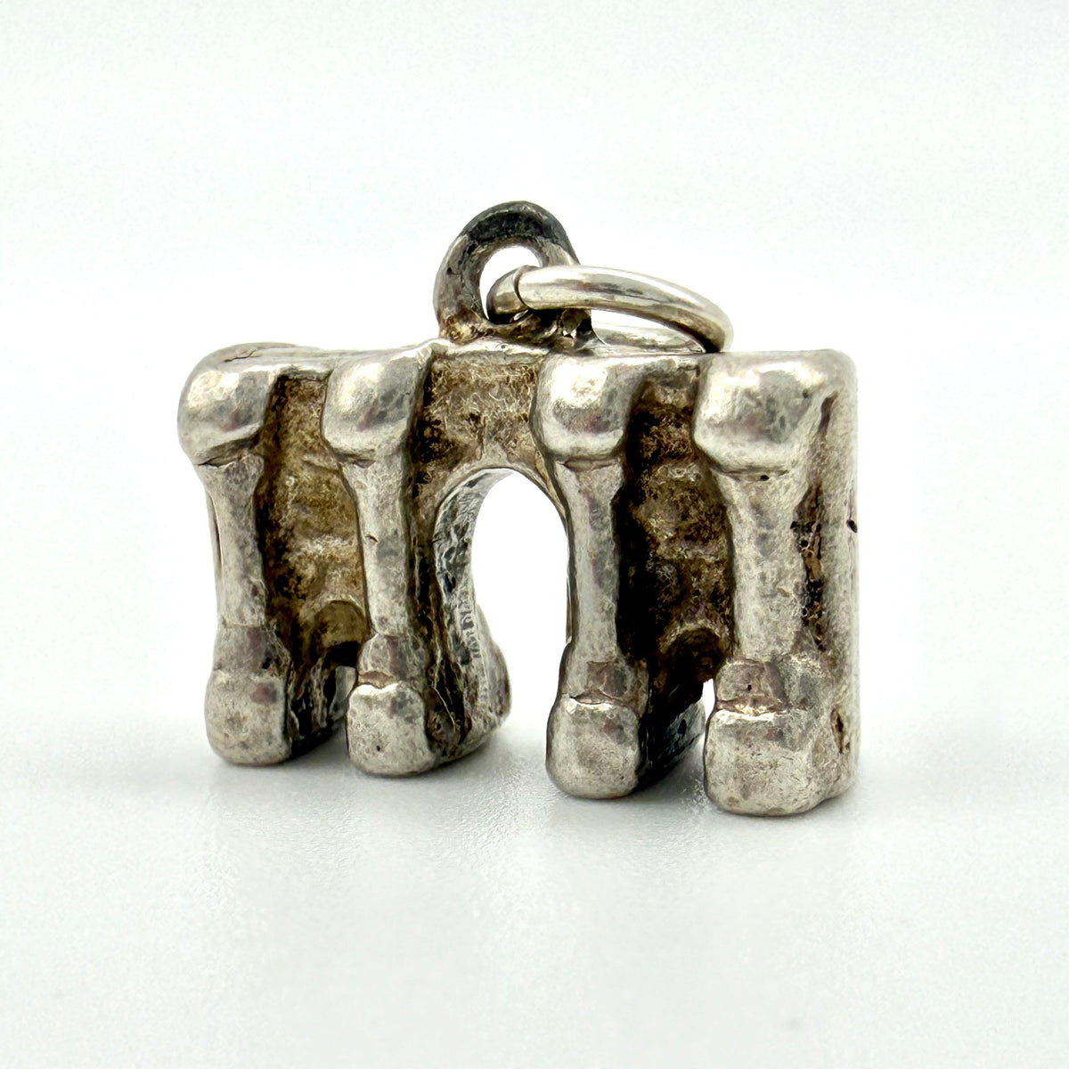 A silver-toned metal charm shaped like a stylized archway with four vertical textured sides and a small top loop, photographed on a plain light background.