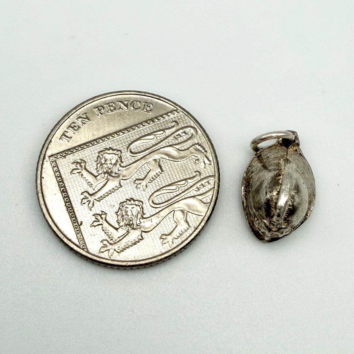 A small silver pendant or charm shown next to a silver ten pence coin for scale, on a plain light background.