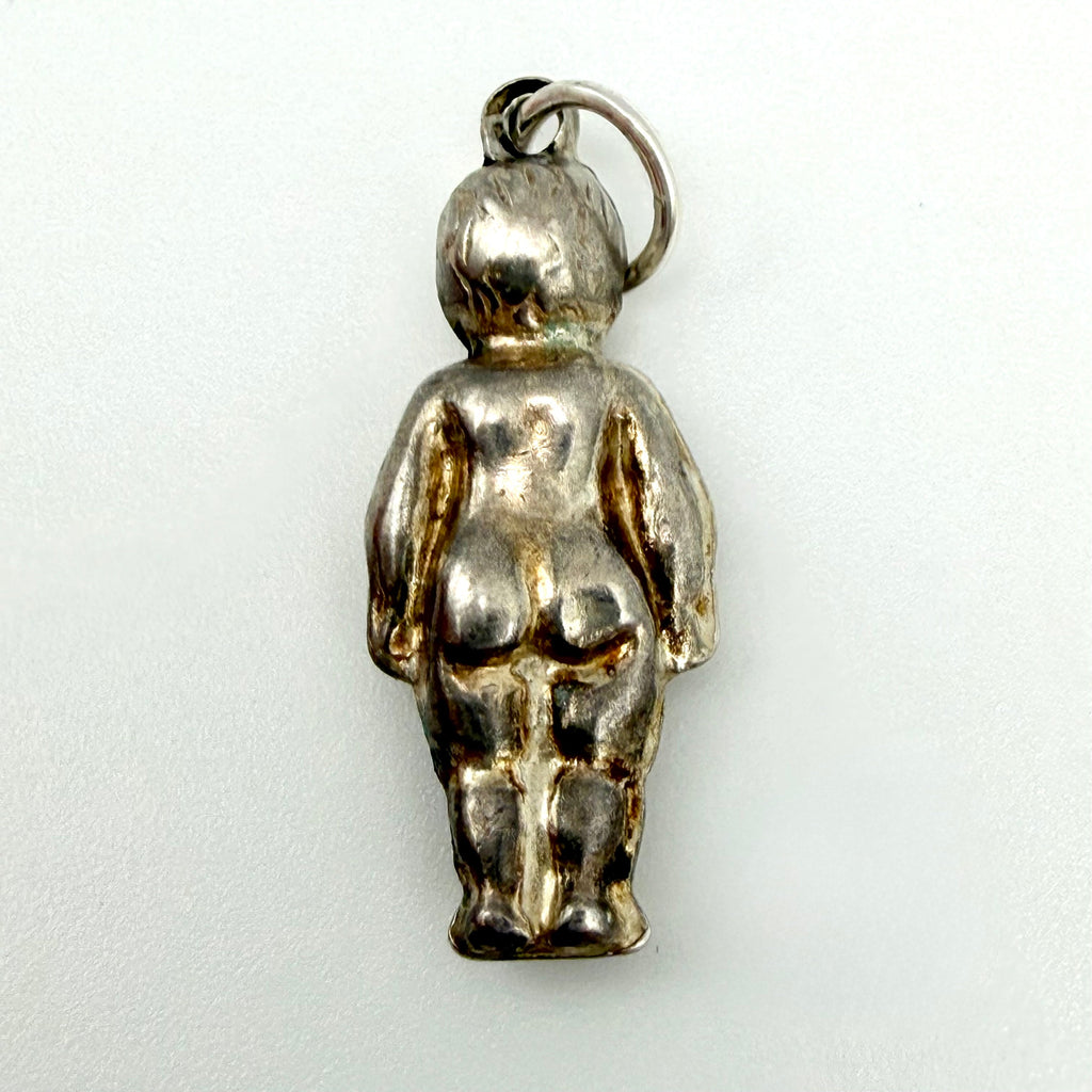 A small metallic pendant shaped like a standing nude figure, photographed against a plain light background. The item appears to be made of silver-toned metal with warm gold-toned highlights from reflections.