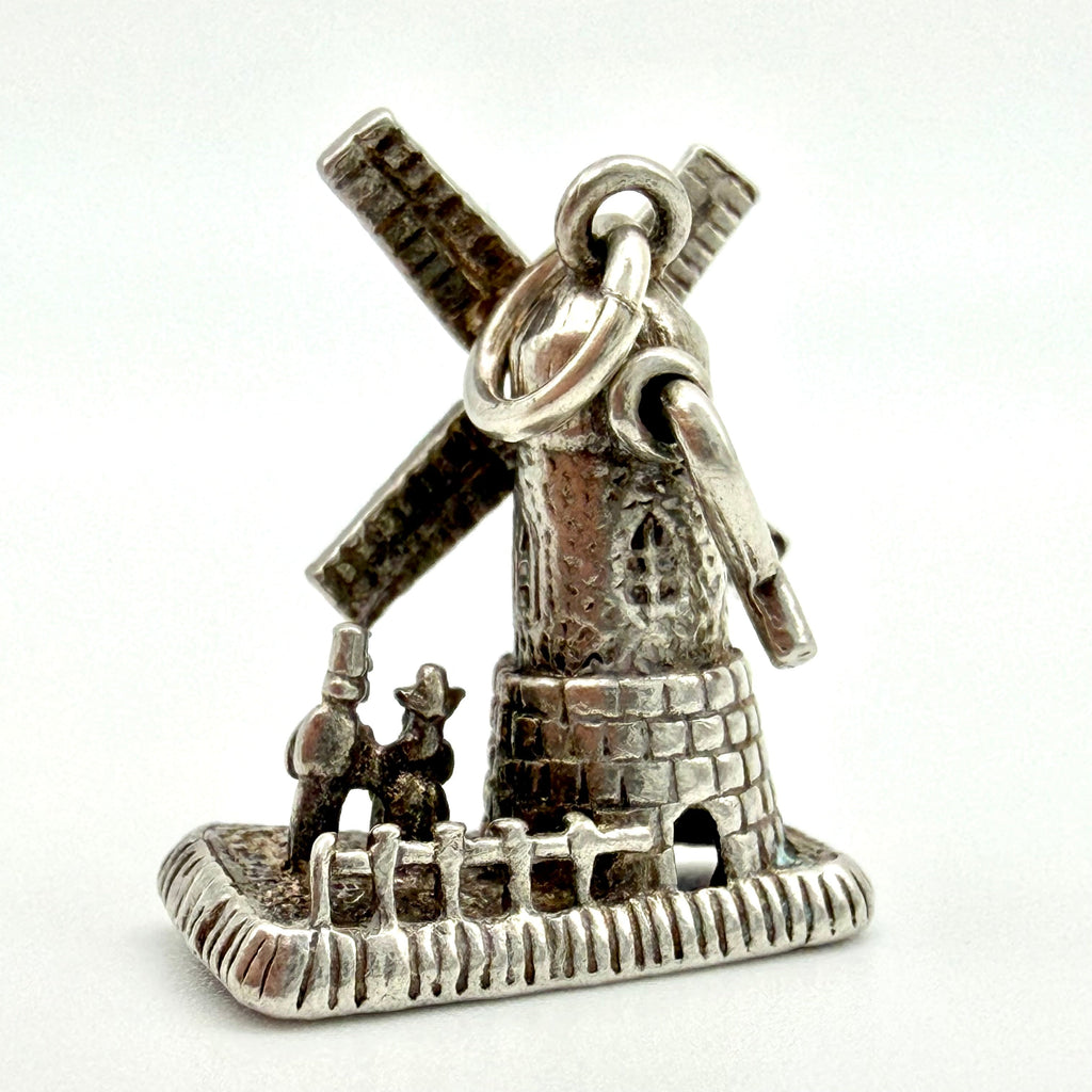 A silver charm shaped like a windmill with two small figures in front, photographed on a plain white background.