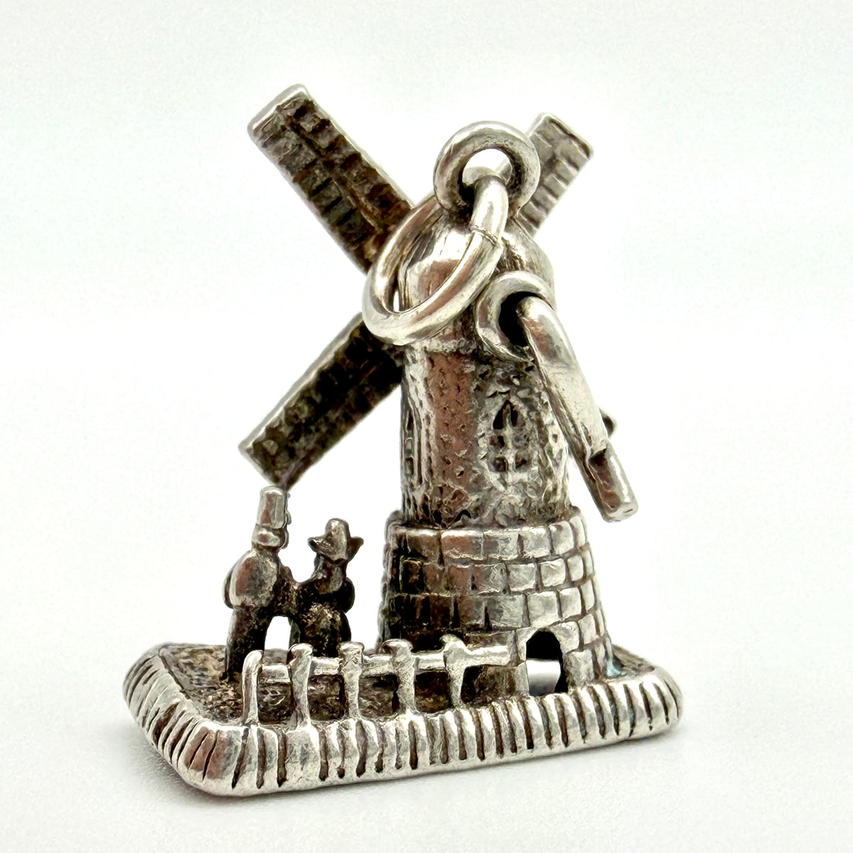A silver charm shaped like a windmill with two small figures in front, photographed on a plain white background.