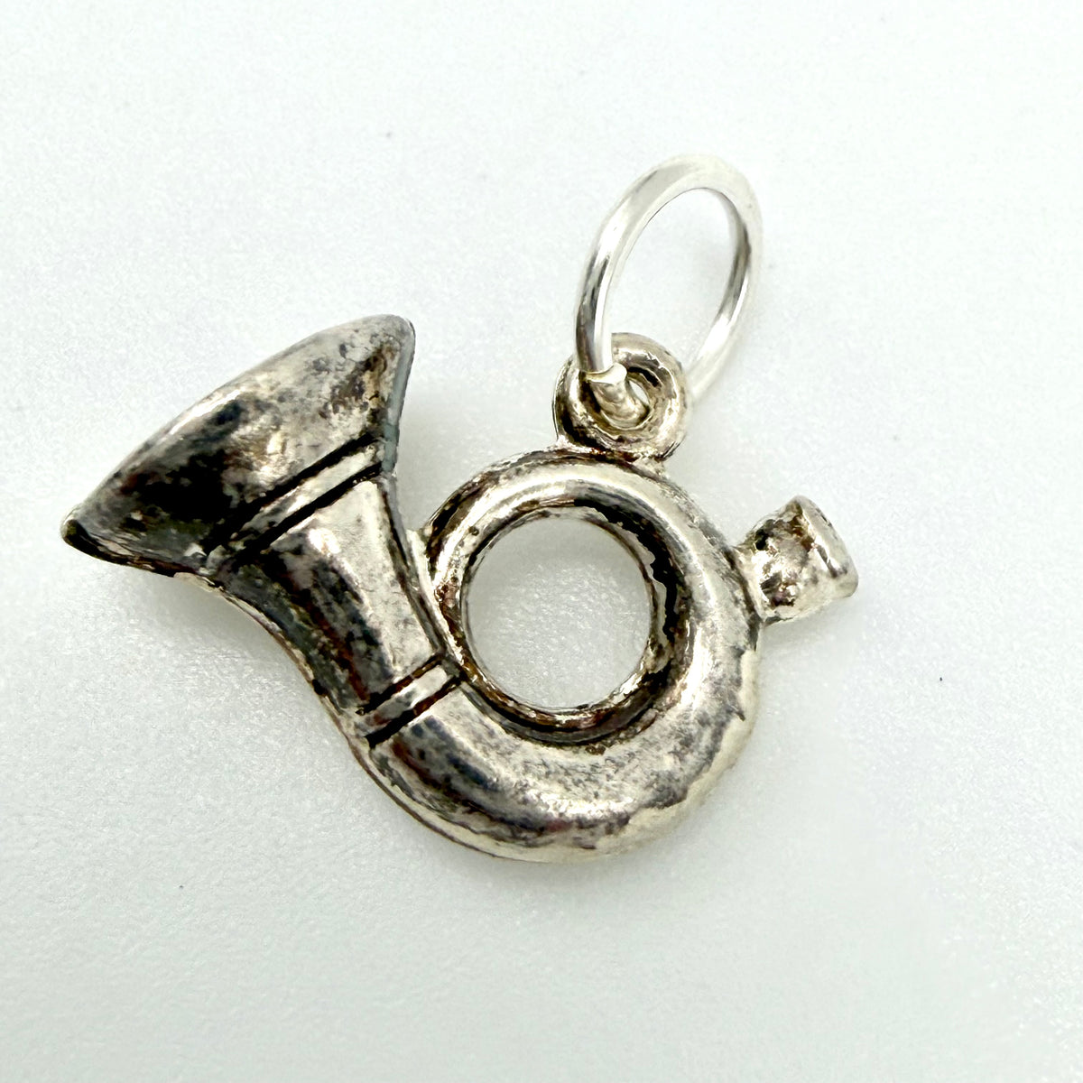A silver metal charm shaped like a French horn, featuring a textured hammered finish and a small hanging loop at the top, shown on a plain white background.