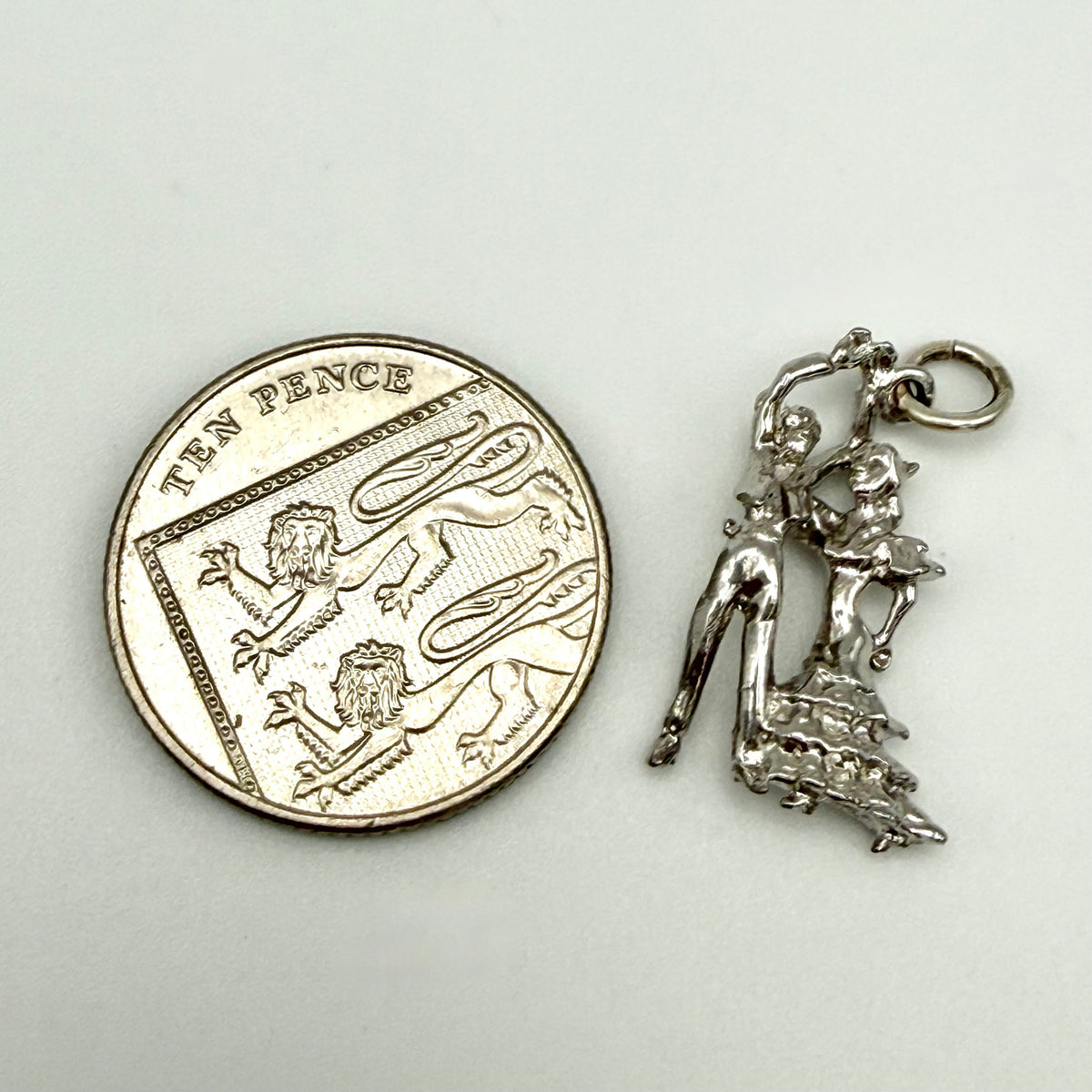A sterling silver charm shaped like a dancing couple, shown next to a ten-pence coin for scale on a plain light background.