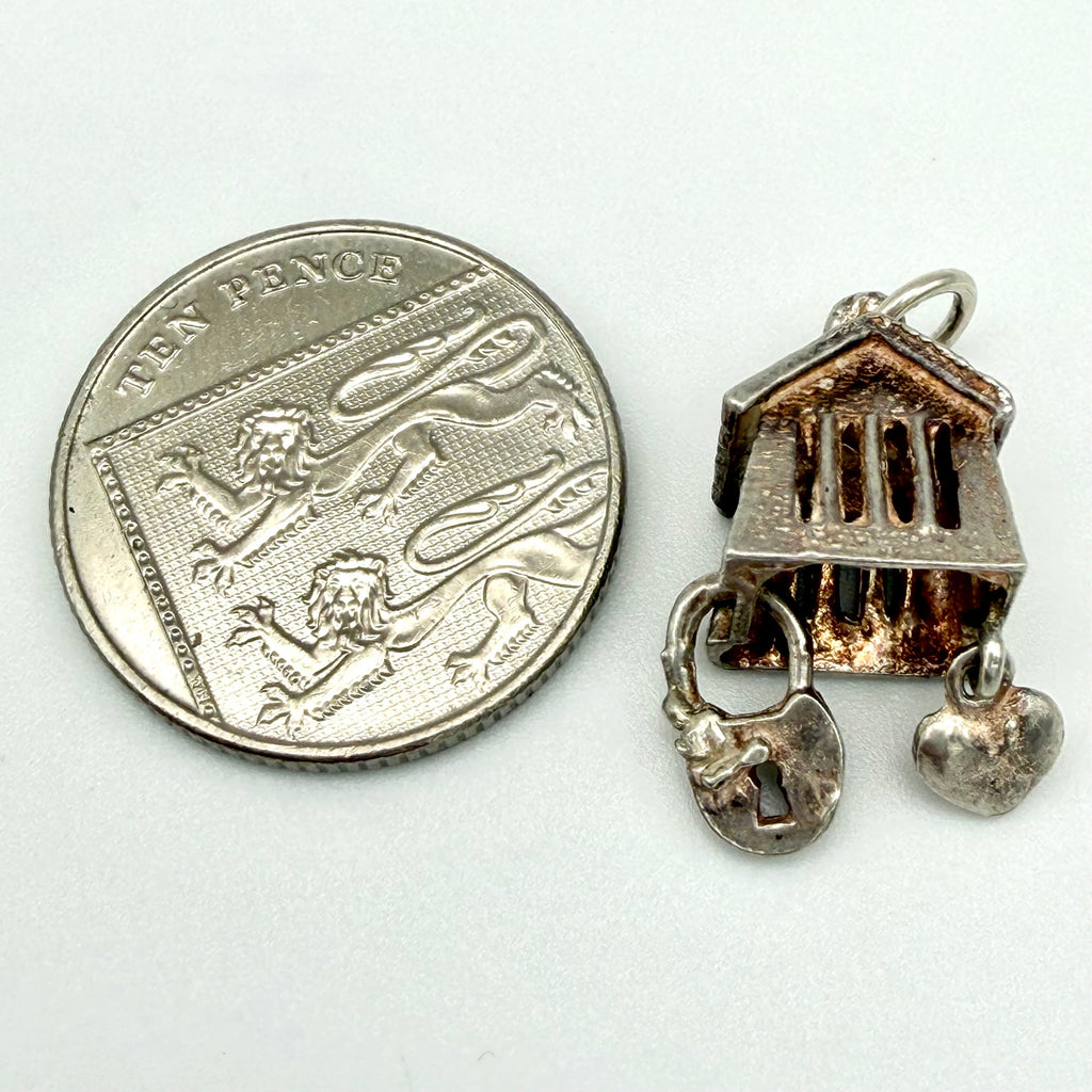 A small silver-toned jewelry charm shaped like a classical building with hanging padlock and heart charms, shown next to a UK ten-pence coin for scale on a plain light background.