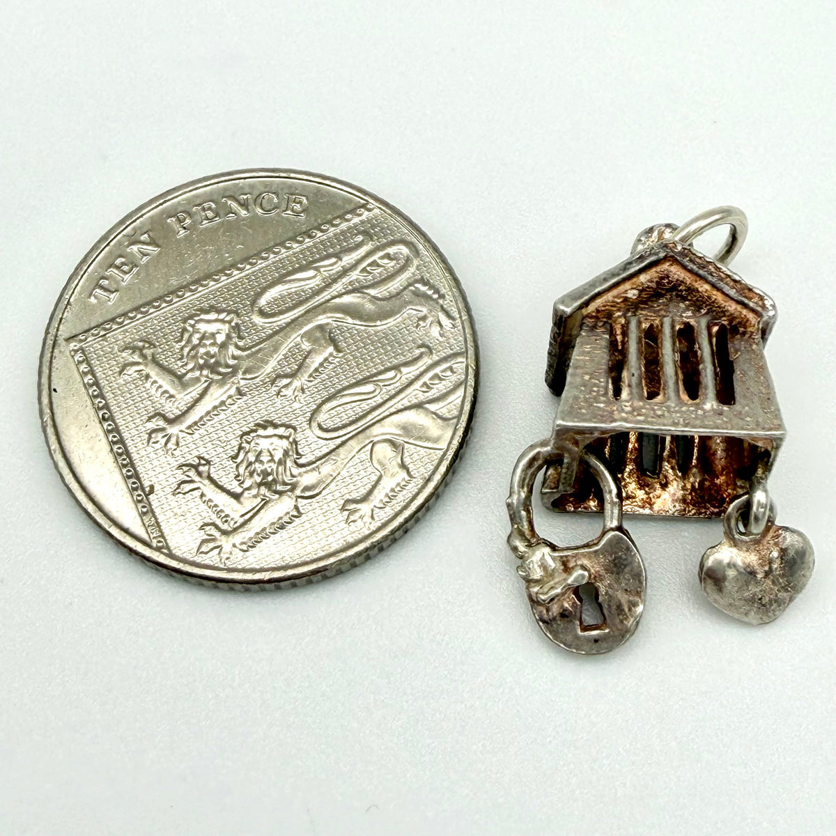 A small silver-toned jewelry charm shaped like a classical building with hanging padlock and heart charms, shown next to a UK ten-pence coin for scale on a plain light background.