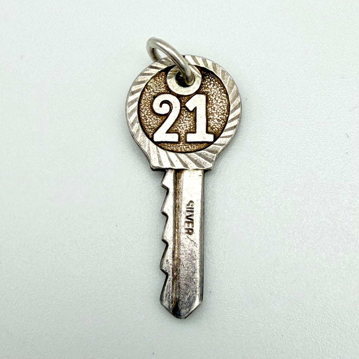 A silver-toned key-shaped pendant charm engraved with the number 21 on the key bow, shown on a plain light gray background.