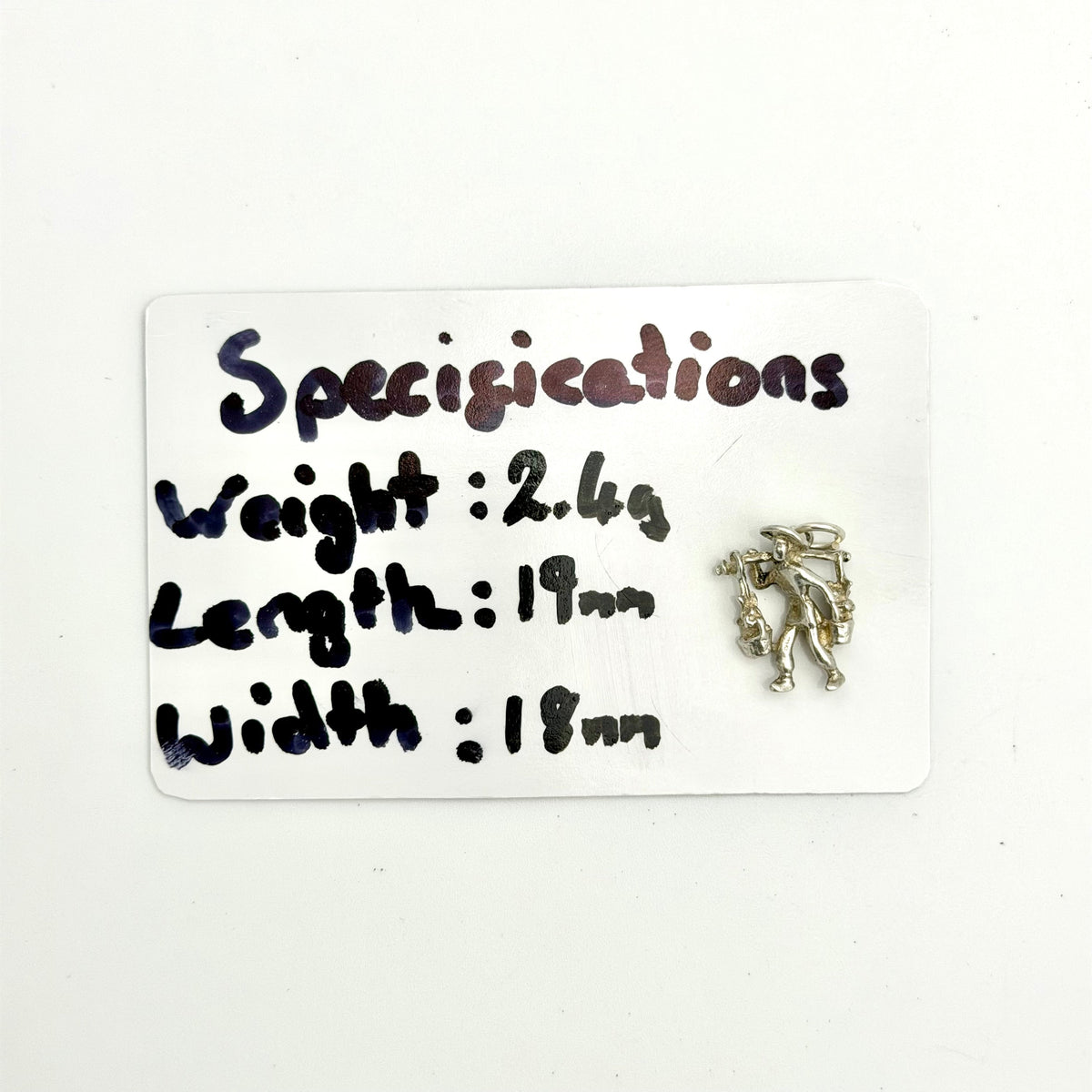 A small silver-toned metal charm or pendant shaped like a stylized figure, placed next to a white handwritten card labeled 'Specifications' with measurements and weight.