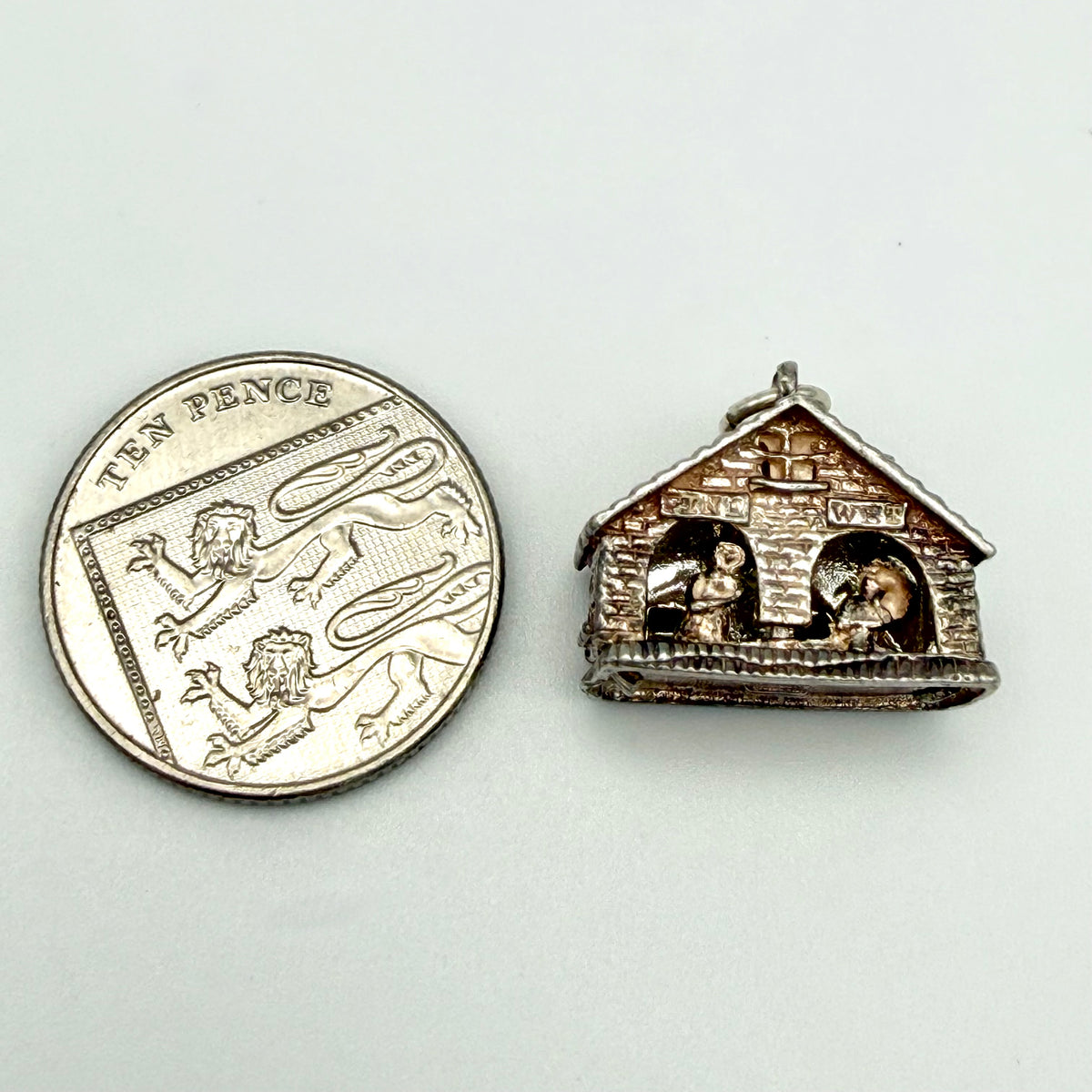 A small silver-toned house-shaped pendant or charm shown next to a ten pence coin for scale, on a plain light background.