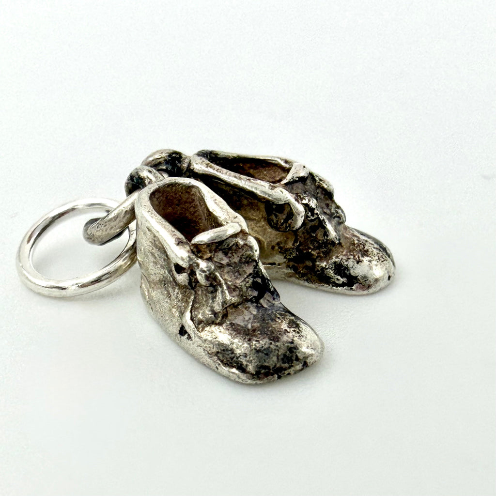 A pair of miniature silver baby boot charms with a distressed, oxidized finish and a small silver keyring attached, shown on a plain white background.