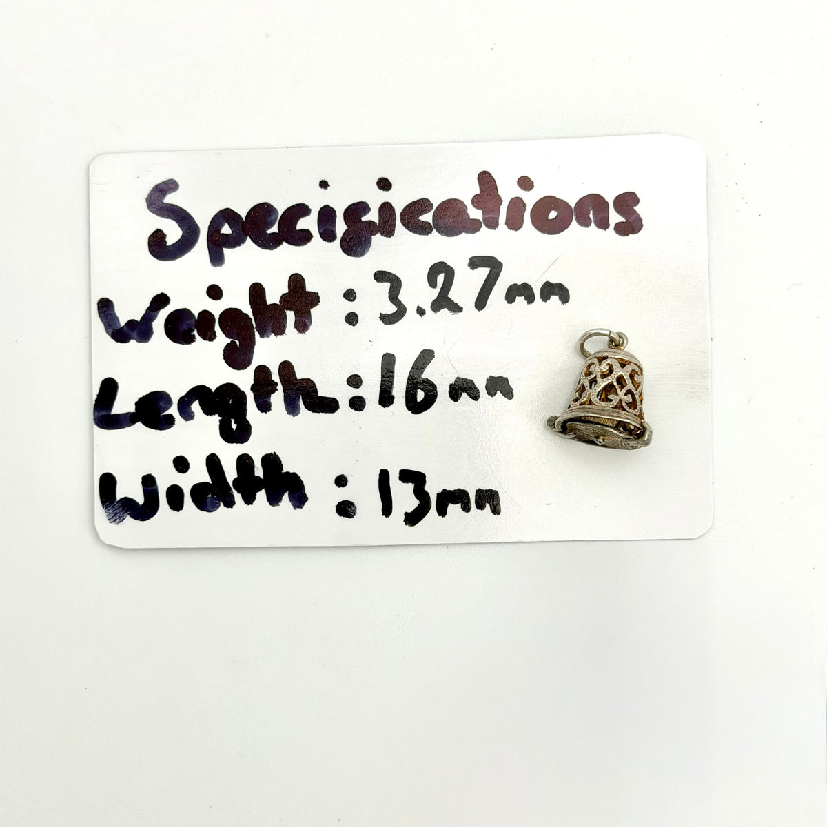 A small silver-toned bell-shaped pendant or charm with a cutout lattice design, placed next to a white card labeled 'Specifications' with handwritten measurements and weight.