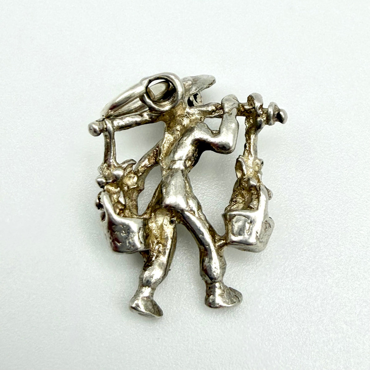 A silver-toned metal pendant or brooch depicting a stylized figure carrying baskets, photographed on a plain light background.