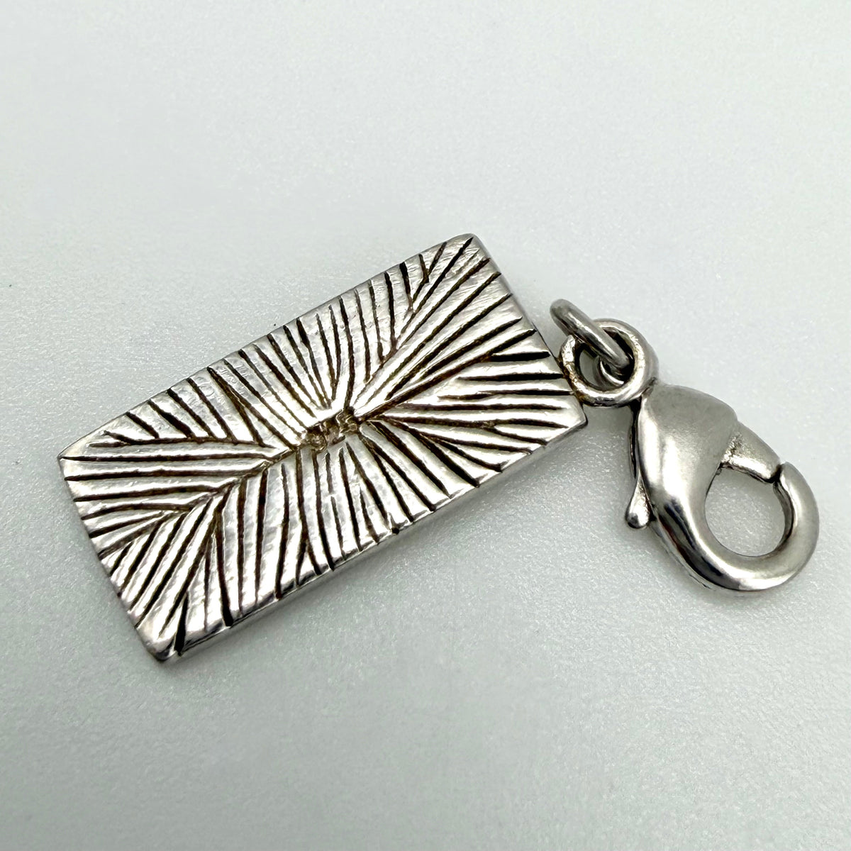 A silver rectangular charm pendant with a textured sunburst or leaf-like engraved pattern, attached to a silver lobster clasp, shown on a light gray background.