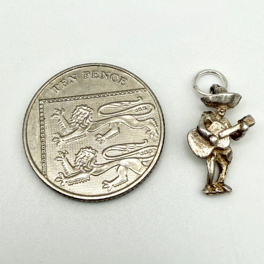A small gold-tone Mexican mariachi player charm shown next to a silver 10 pence coin for scale, on a plain light background.