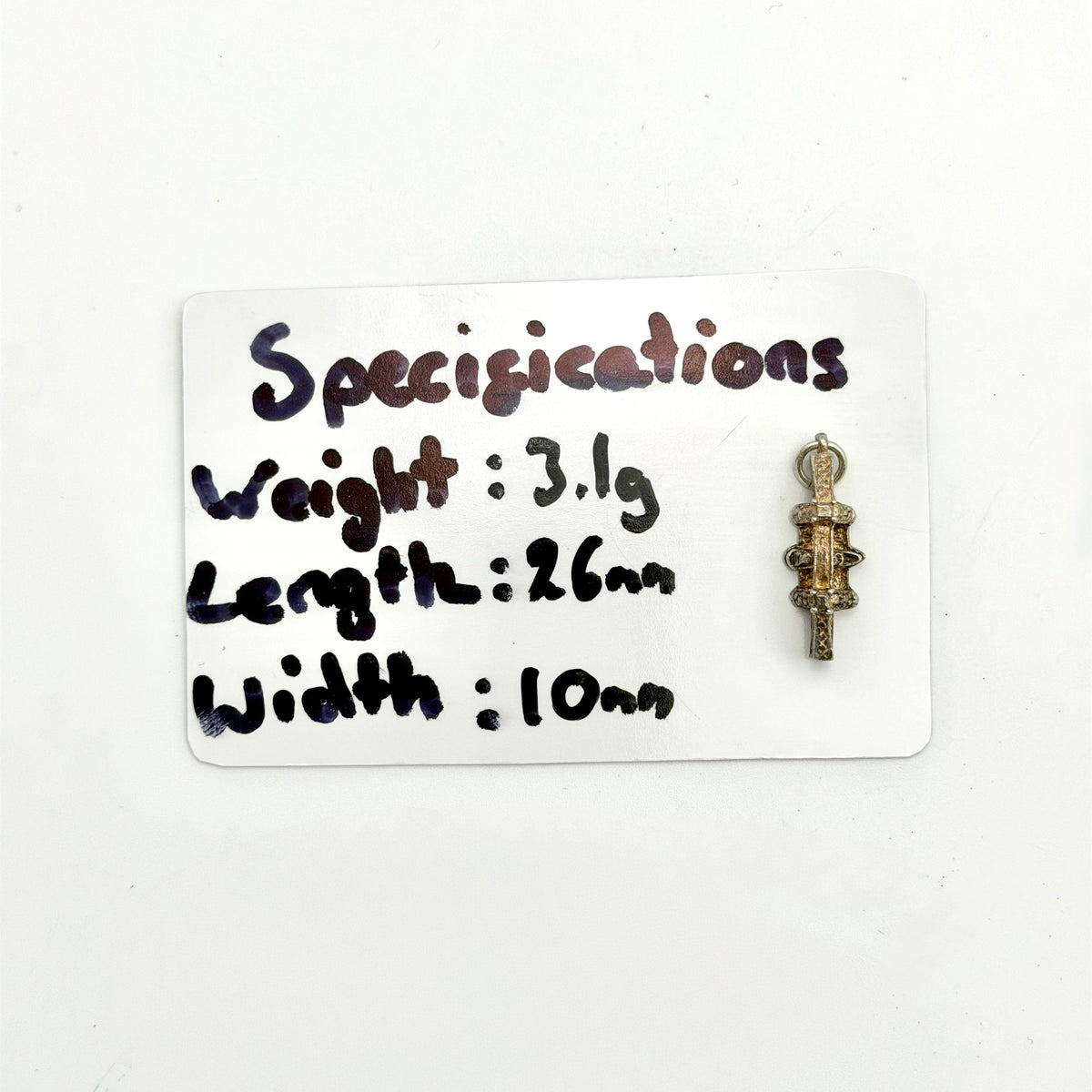 A small antique-style metal pendant or charm displayed next to a white handwritten card showing its specifications: weight 3.1g, length 26mm, and width 10mm.