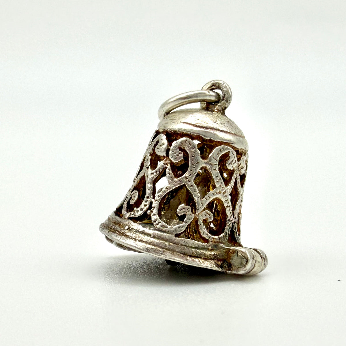 A small silver bell-shaped pendant charm with intricate filigree cutouts and a top loop, photographed on a plain light background.
