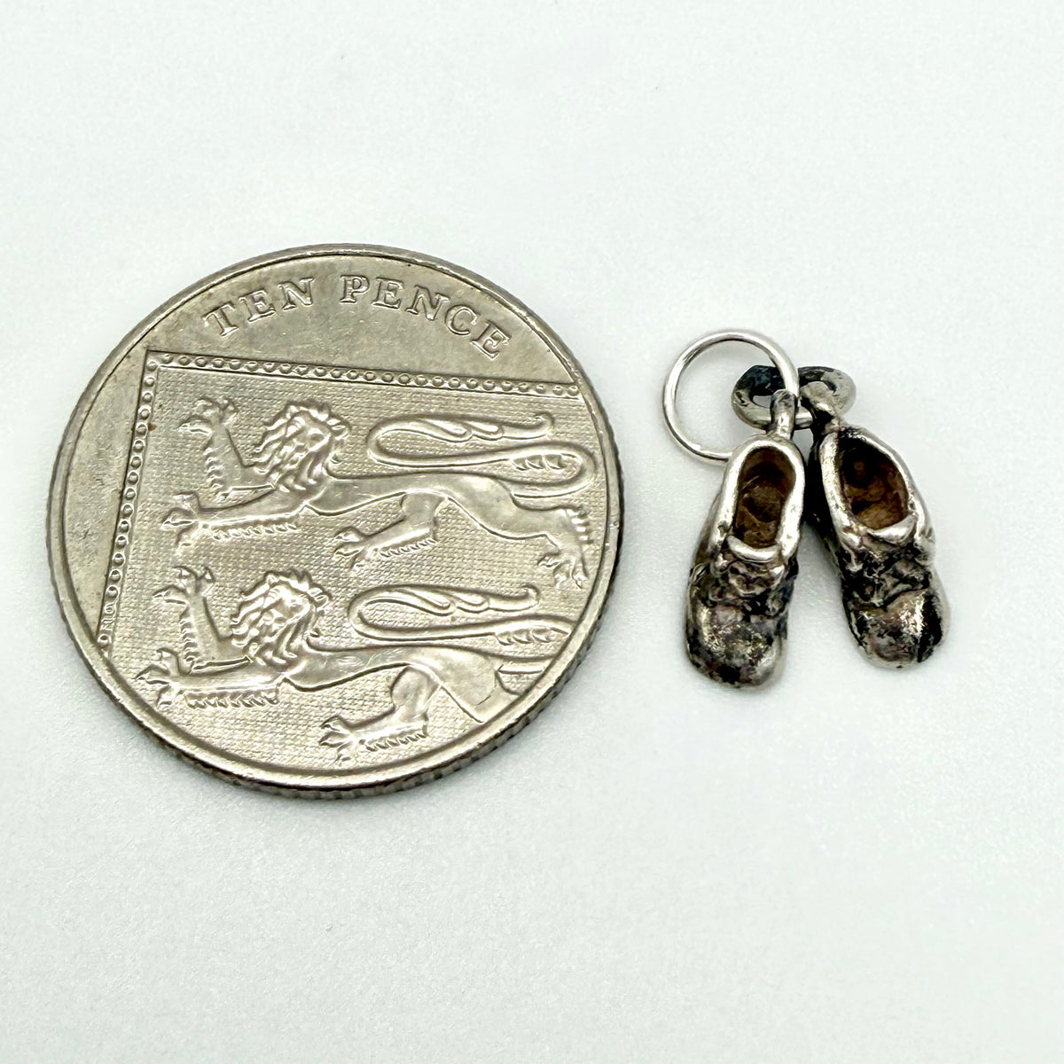 A pair of miniature silver Dutch shoes charm earrings shown next to a ten pence coin for scale, on a plain white background.