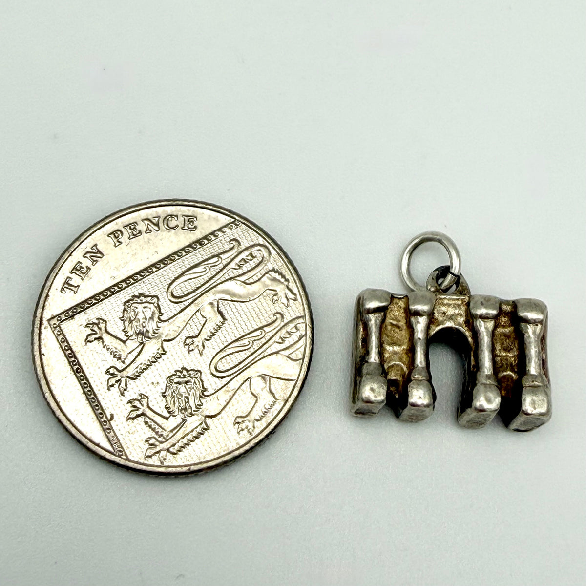 A small silver-toned metal charm shaped like three vertical teeth with a ring at the top, shown next to a UK ten pence coin for scale on a light gray background.