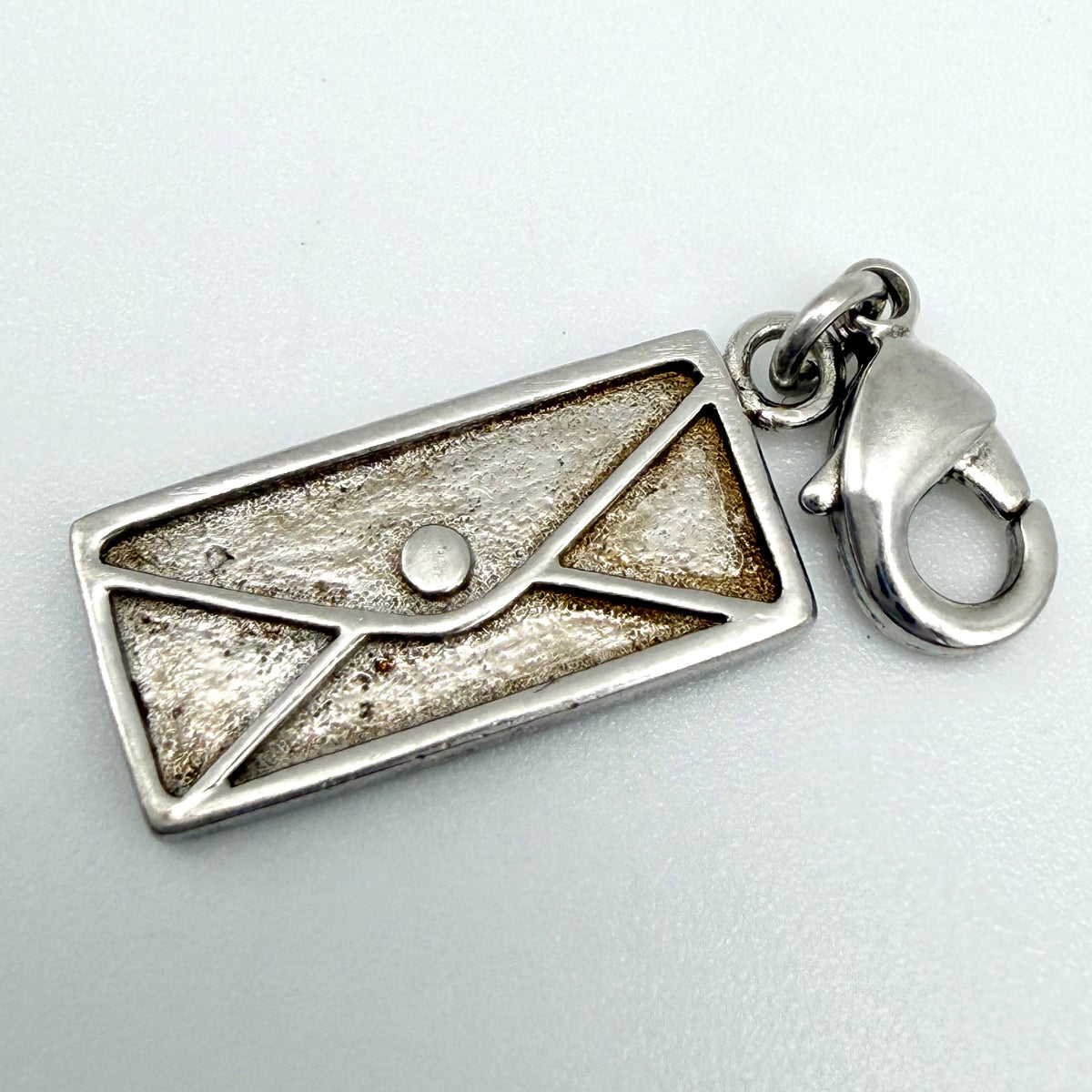 A silver metal charm shaped like an envelope with a textured, antiqued center and a lobster clasp attached on the right side, shown on a light gray background.