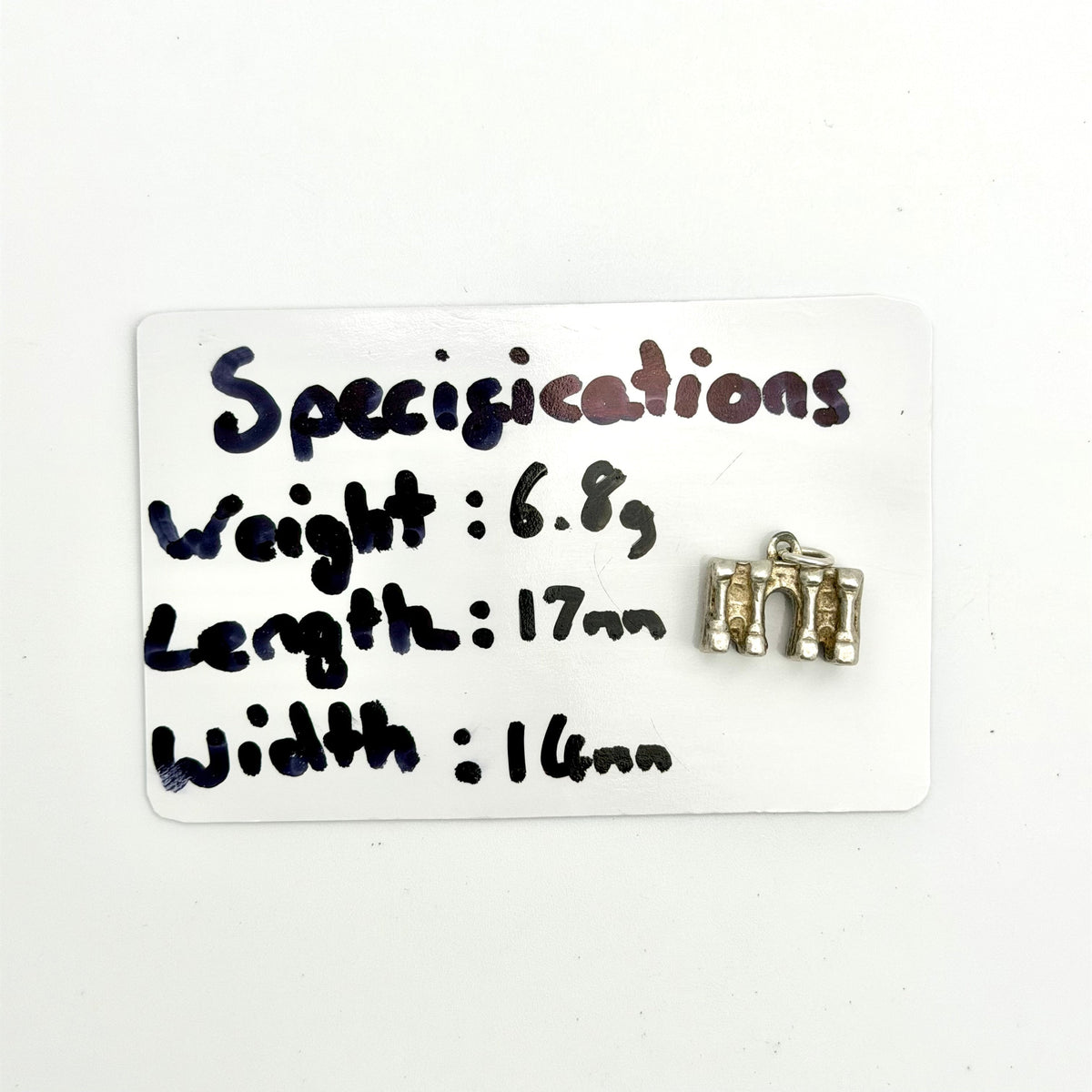 A small silver-toned metal jewelry piece, likely a charm or pendant, displayed on a white card labeled 'Specifications' with handwritten measurements and weight.