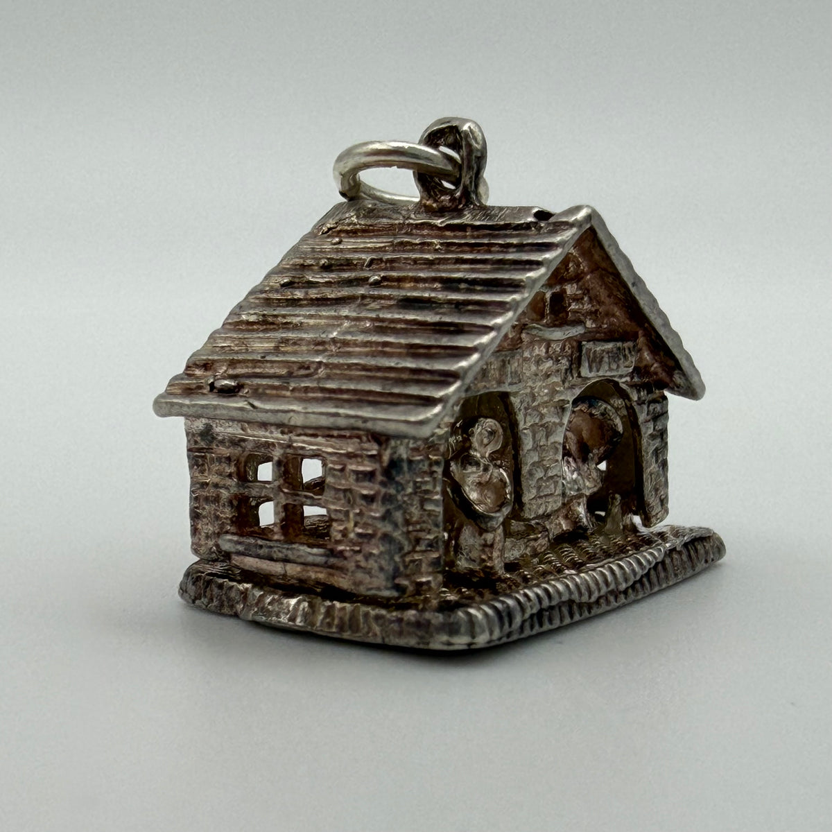 A small silver house-shaped pendant charm with a textured finish and a hanging loop at the top, photographed on a plain light gray background.
