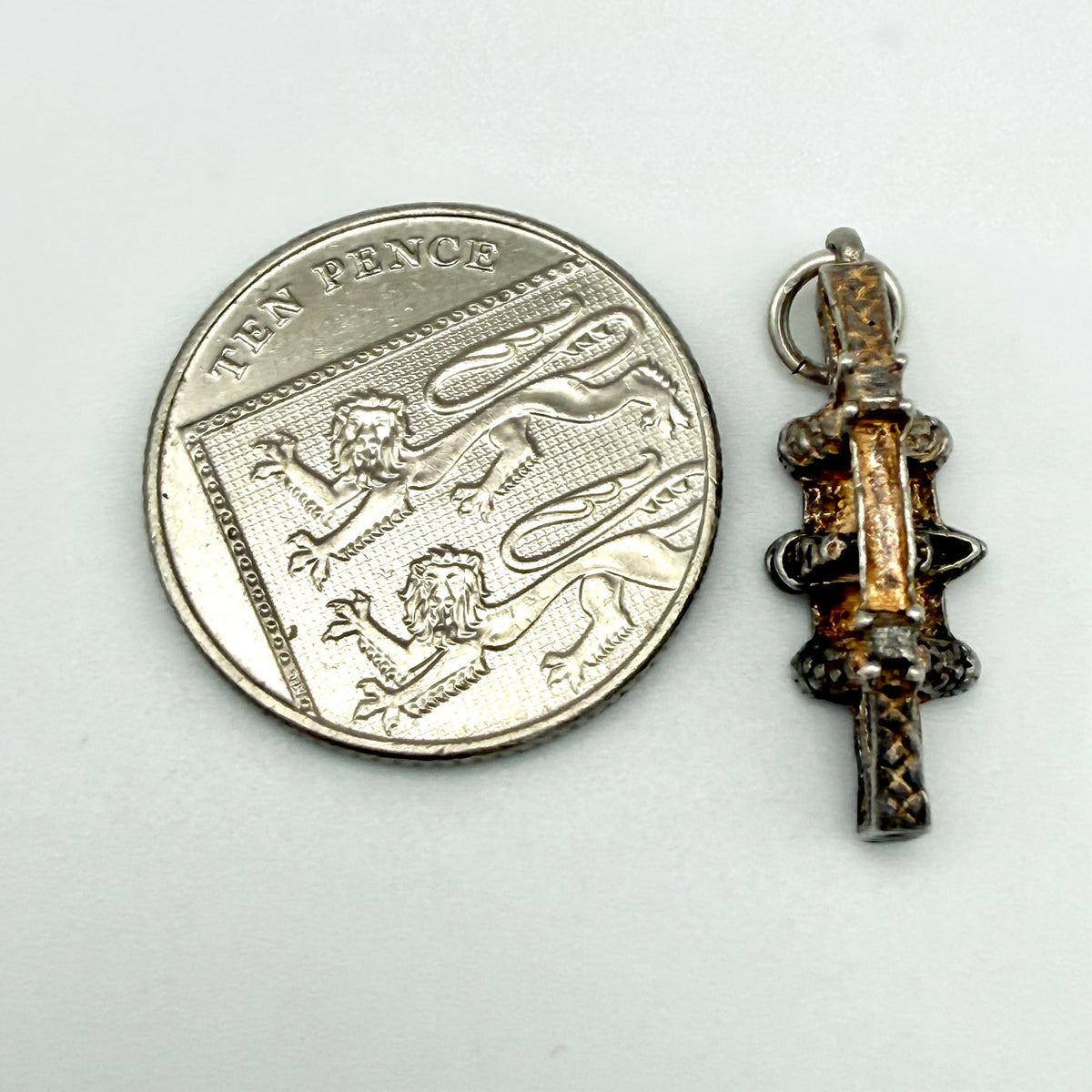 A small ornate pendant or charm shown next to a silver ten pence coin for scale, on a plain light background.