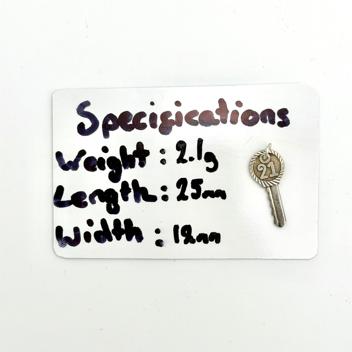 A small silver-toned key-shaped accessory displayed next to a white card with handwritten specifications listing weight, length, and width.