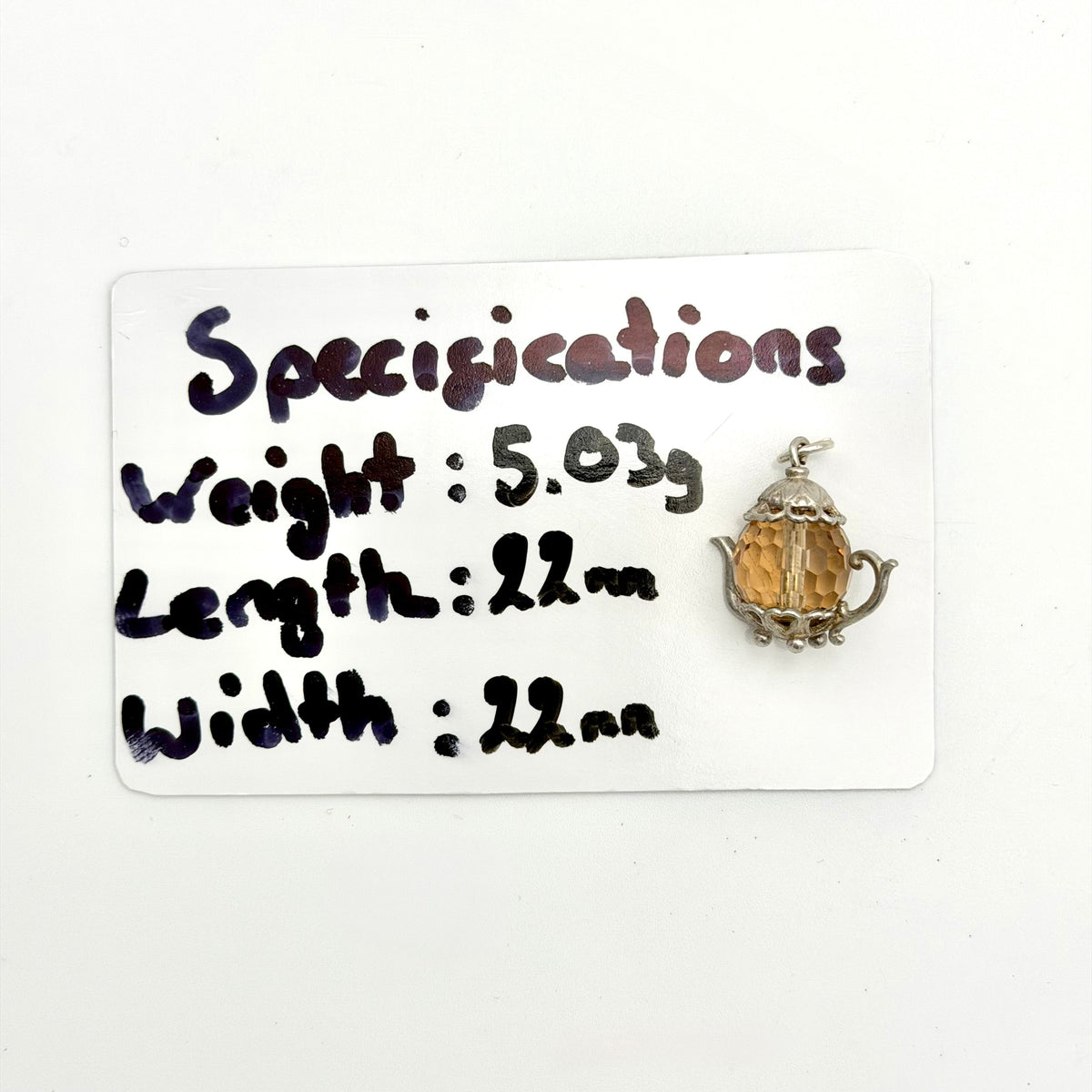 A small silver-toned pendant with a faceted amber-orange center, shown next to a white handwritten specification card that lists weight, length, and width.