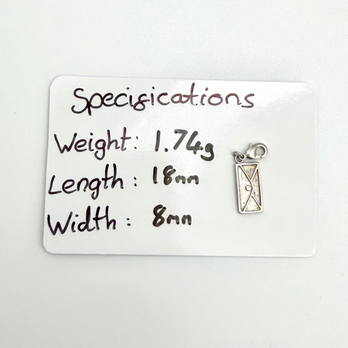 A small silver-toned rectangular pendant or charm with a loop at the top, shown next to a white card labeled 'Specifications' with handwritten measurements and weight.