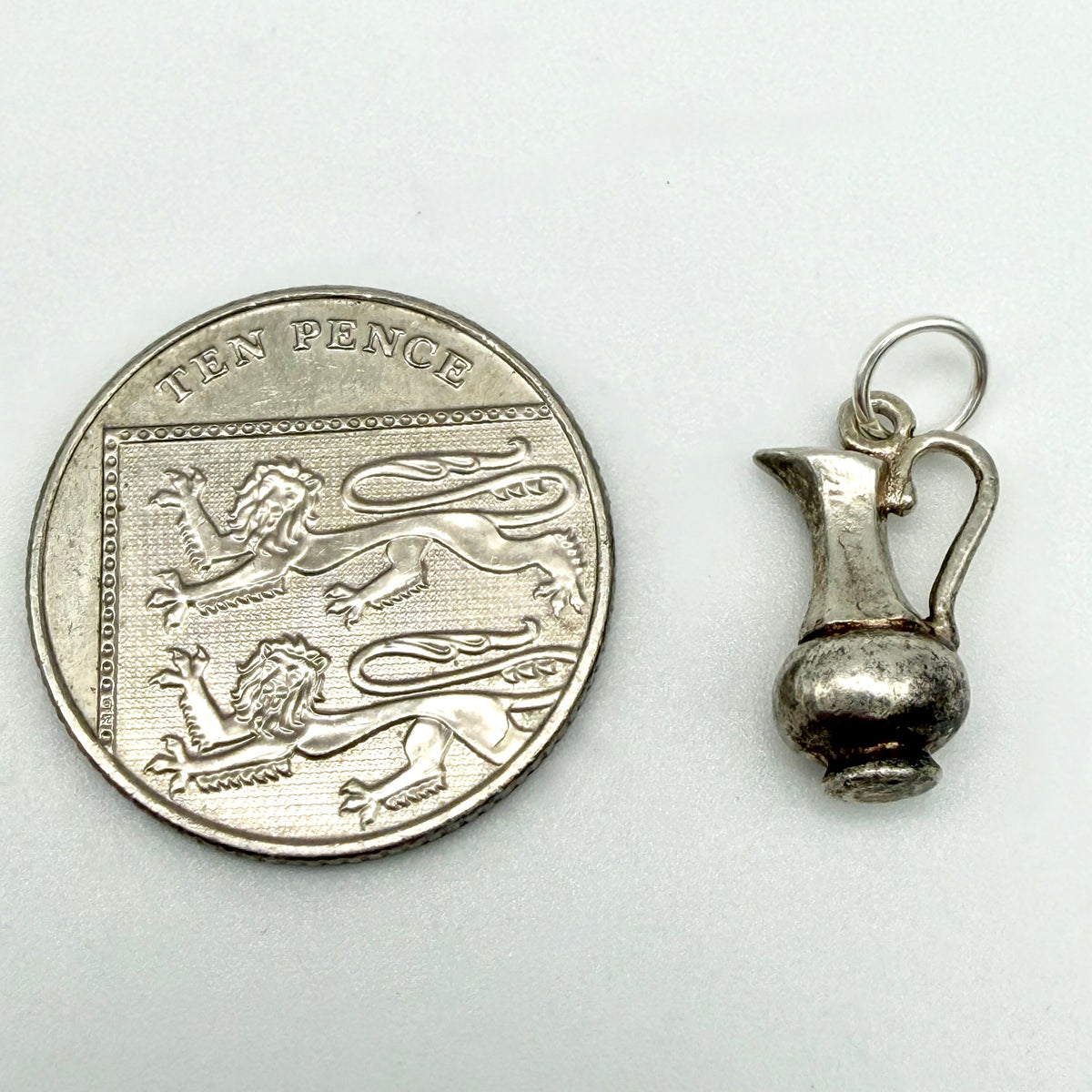 A small silver pitcher charm shown next to a UK ten pence coin for scale, on a plain light background.