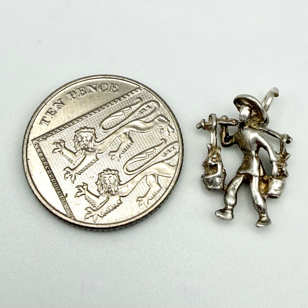 A sterling silver charm of a farmer carrying sheep on his shoulders, shown next to a ten-pence coin for scale on a light gray background.