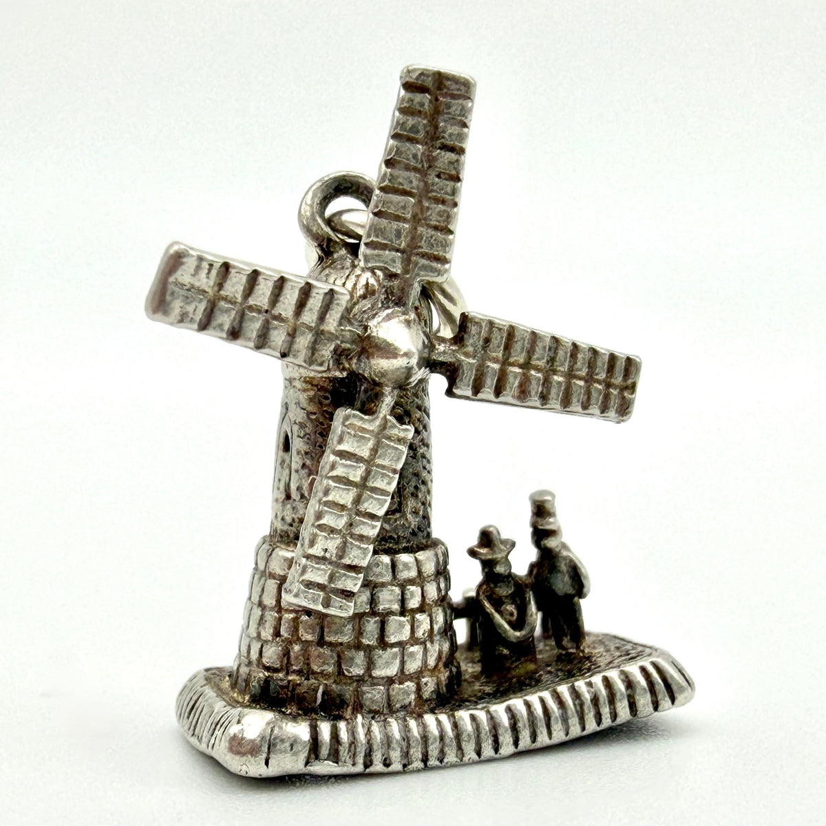 A small silver windmill charm or pendant with two tiny human figures at the base, photographed against a plain light background.