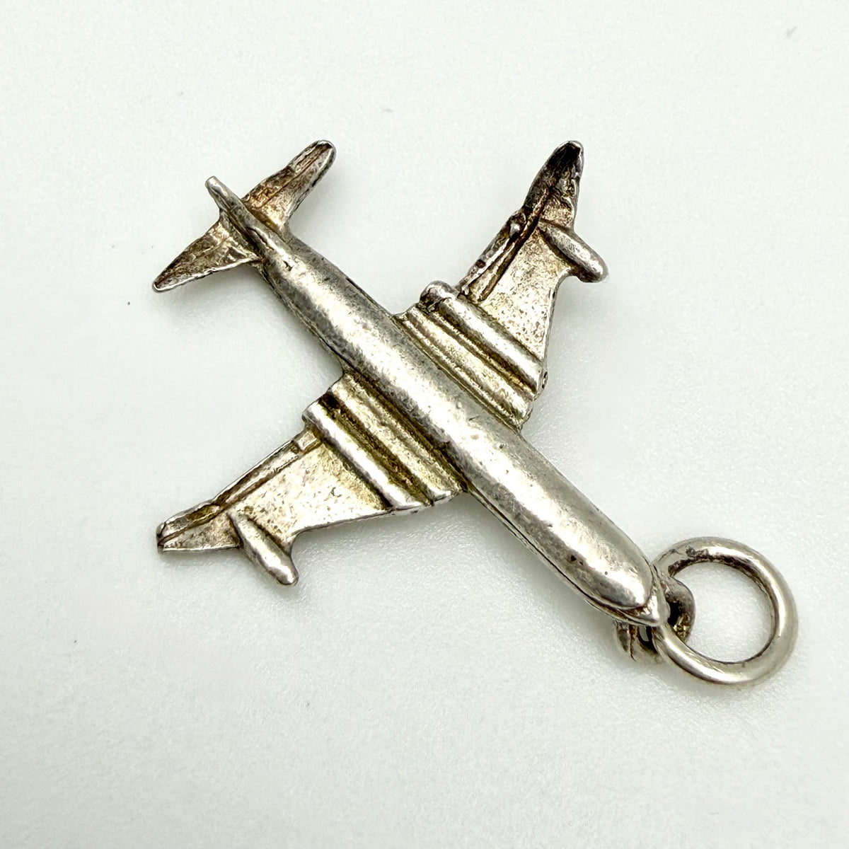 A silver airplane charm pendant with a textured, antiqued finish and a hanging loop at the tail, shown on a plain white background.