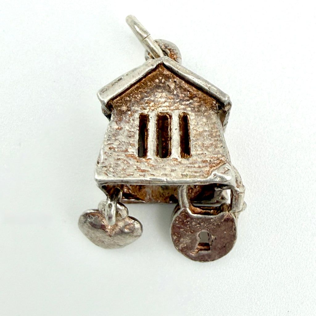 A small house-shaped pendant charm in textured silver-tone metal with a lock and key motif, photographed on a plain white background.