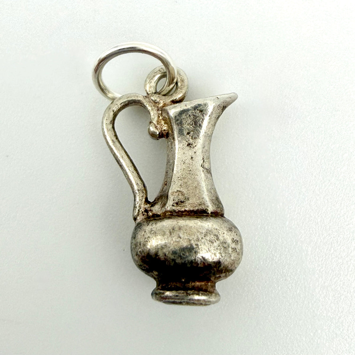 A small silver pitcher charm with a textured, antique finish and a hanging loop, shown on a plain light background.