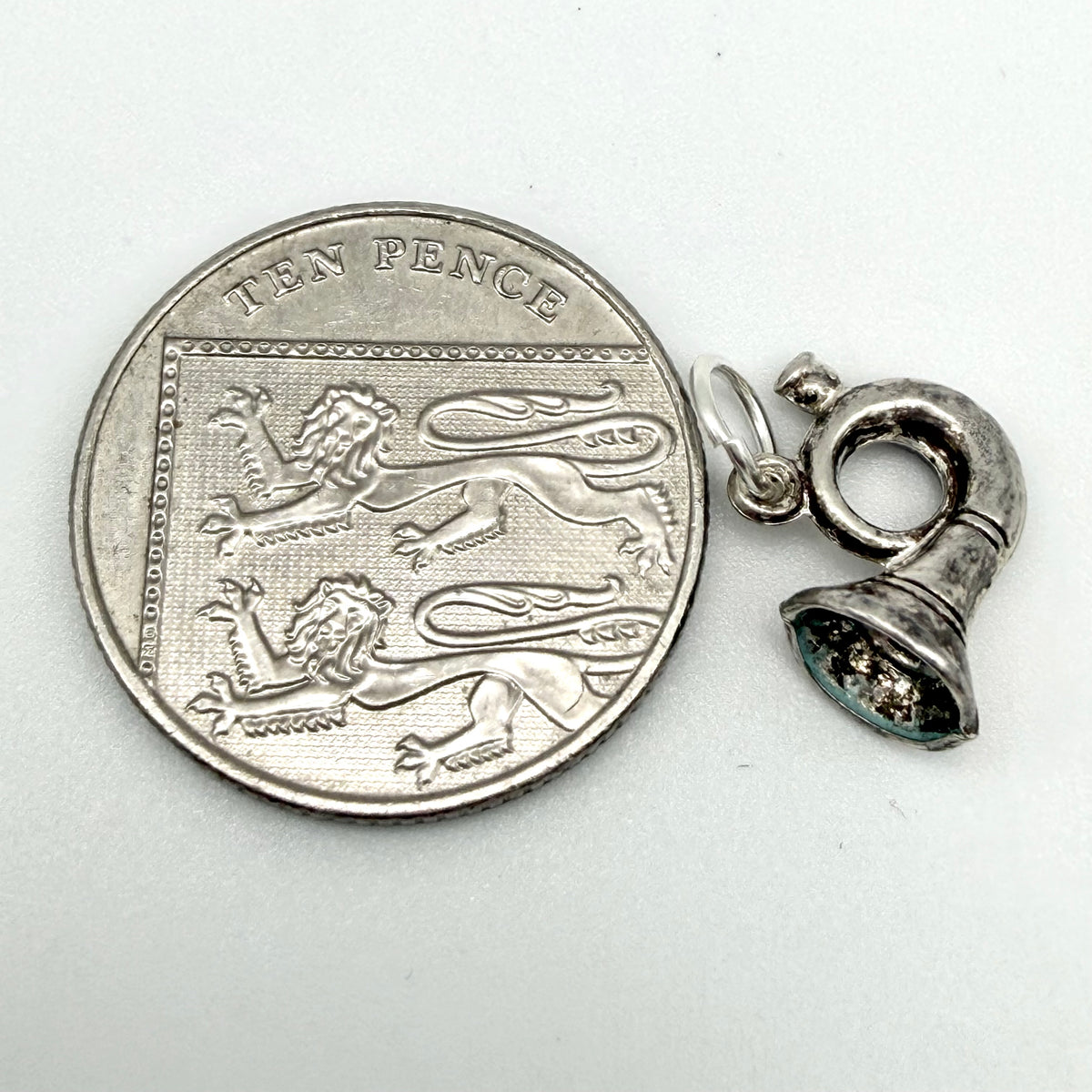 A silver bell-shaped pendant charm shown next to a ten pence coin for scale, on a plain light gray background.