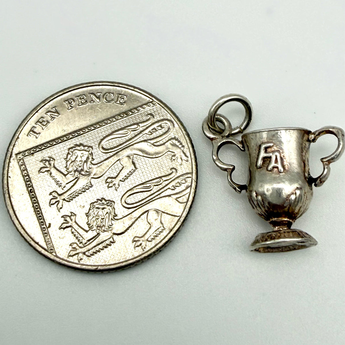 A small silver trophy cup pendant shown next to a UK ten pence coin for scale, on a plain light background.