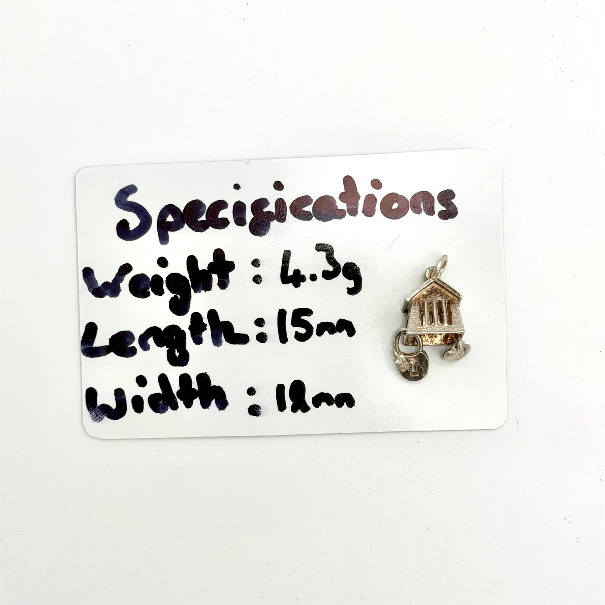 A small jewelry charm shaped like a house on a cart, shown next to a white card with handwritten specifications listing its weight, length, and width.