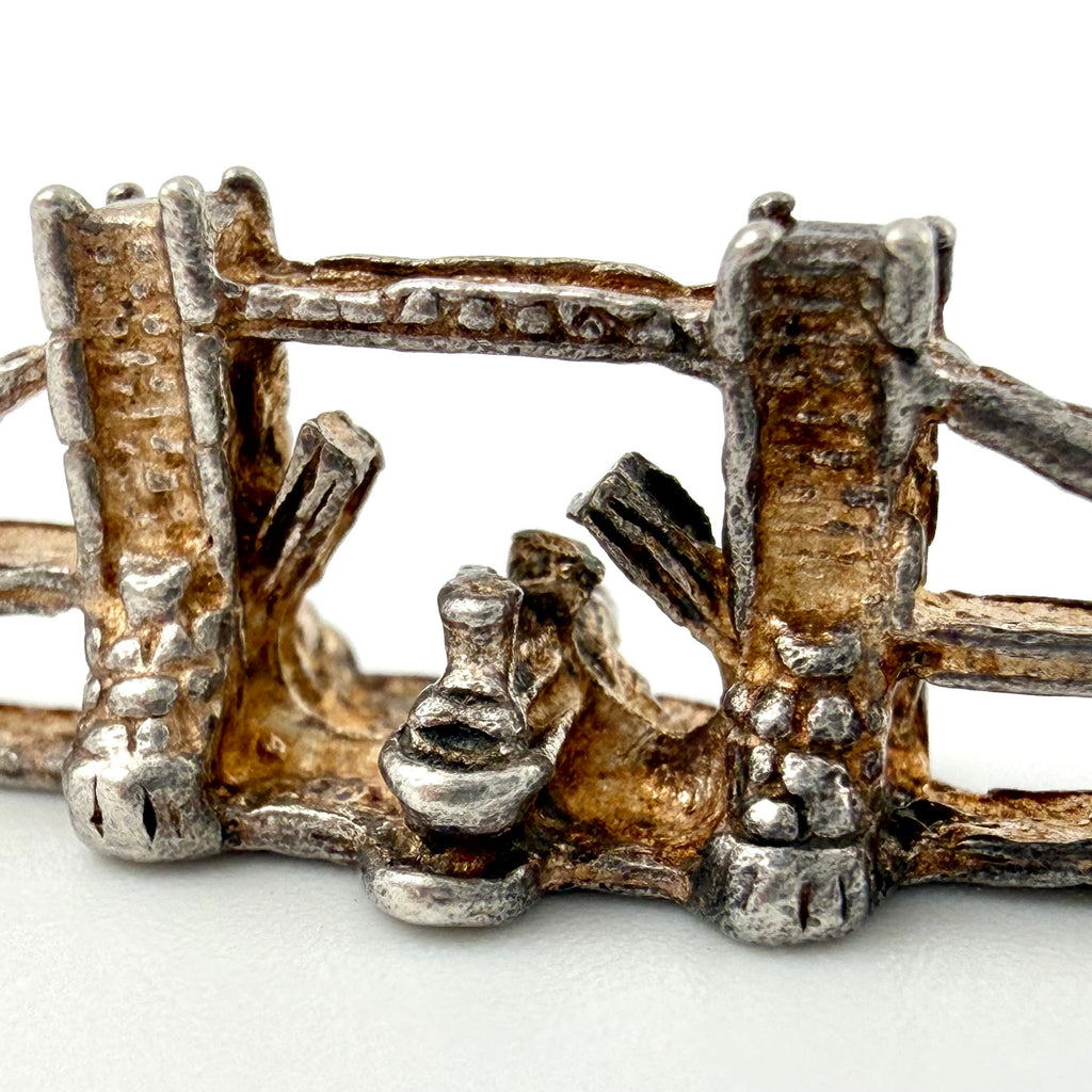 A close-up of a small metallic jewelry piece, likely a miniature charm, brooch, or decorative accent, featuring a rustic, oxidized silver and gold-toned design with textured vertical bars and a central loop.