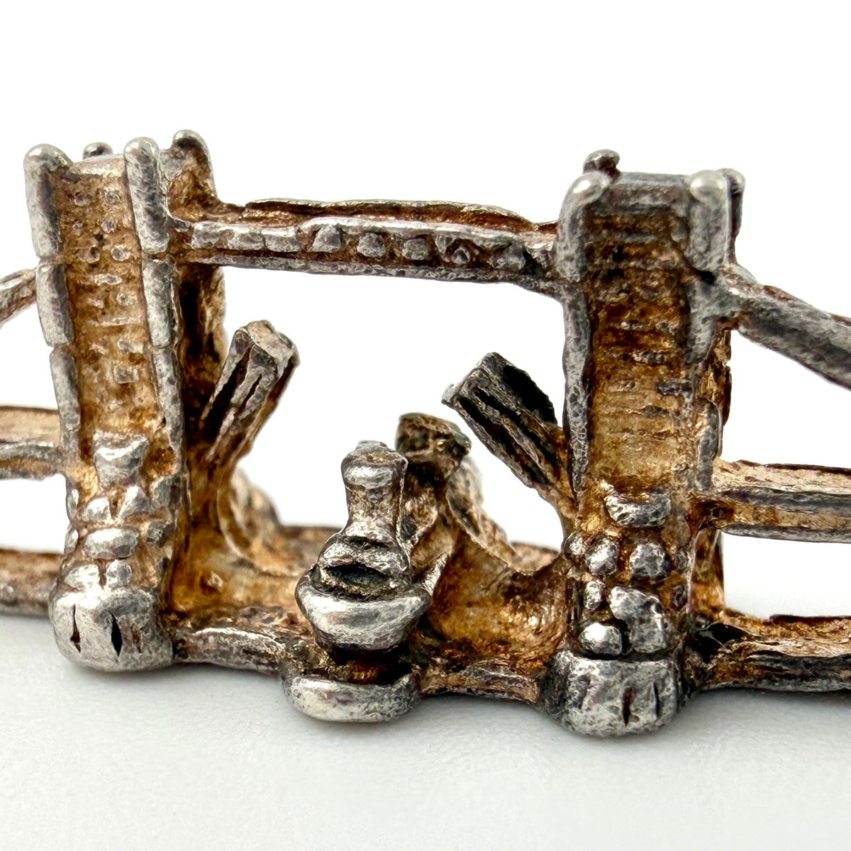A close-up of a small metallic jewelry piece, likely a miniature charm, brooch, or decorative accent, featuring a rustic, oxidized silver and gold-toned design with textured vertical bars and a central loop.