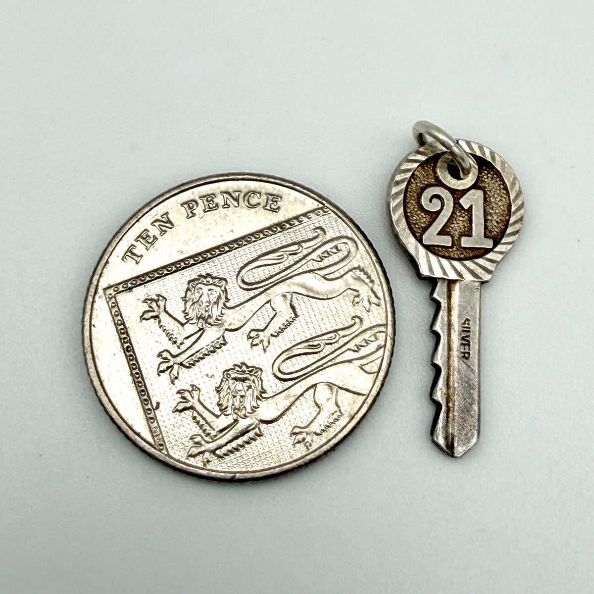 A silver key-shaped pendant charm engraved with the number 21, shown next to a UK ten-pence coin for scale on a light gray background.