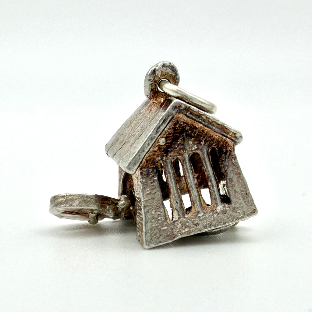A small house-shaped pendant charm with a textured silver finish and a loop at the top, photographed on a plain light background.