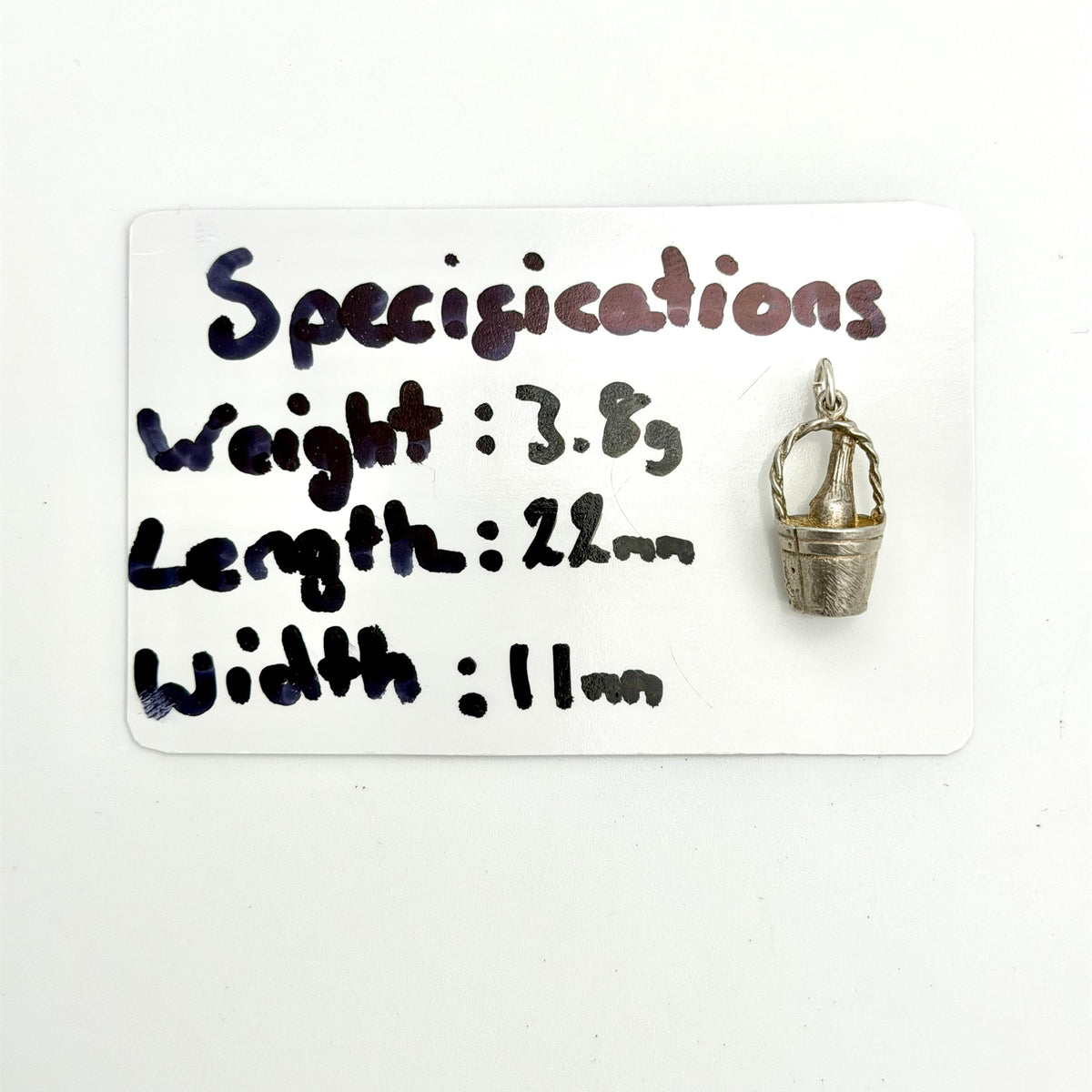 A small silver-toned metal bucket charm is shown next to a white card labeled 'Specifications' with handwritten measurements, weight, and dimensions.