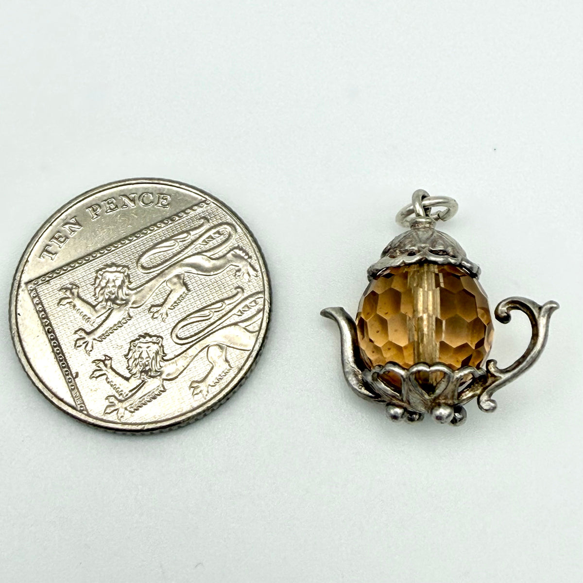 A small silver teapot-shaped pendant with a faceted amber-yellow crystal center, shown next to a silver ten-pence coin for scale on a plain white background.