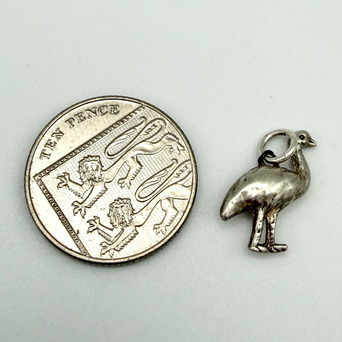 A small silver bird charm, shaped like a pheasant, shown next to a UK ten pence coin for scale on a light gray background.